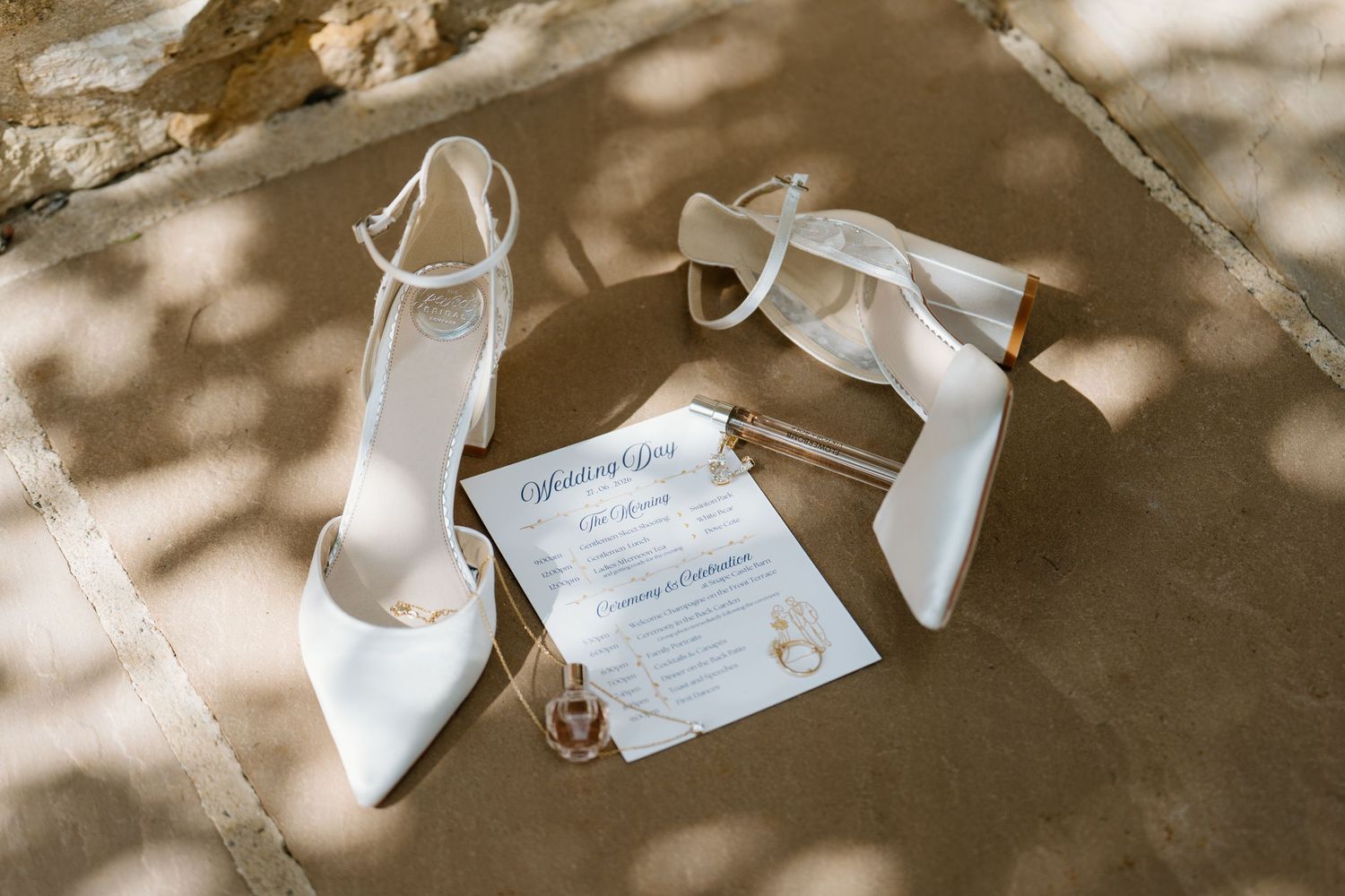 White satin bridal heels and wedding invitation displayed on stone surface with soft shadows.