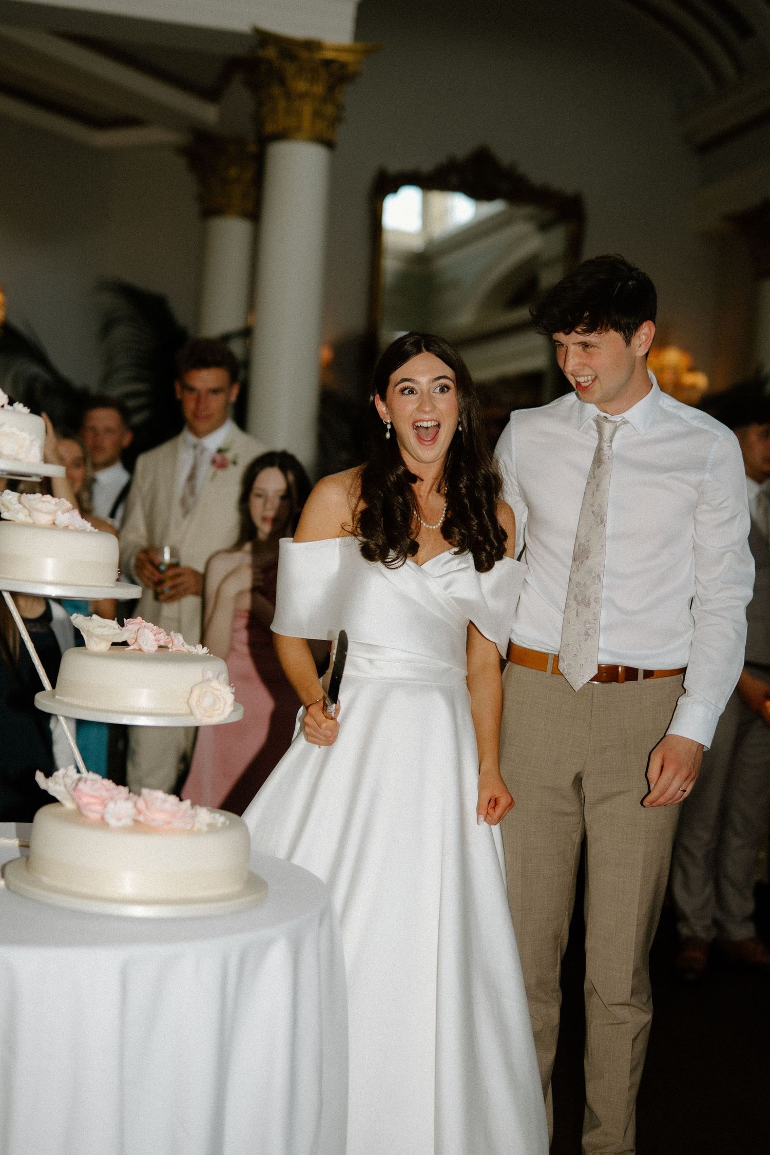 Excited bride and groom preparing to cut their elegant white tiered wedding cake at reception.