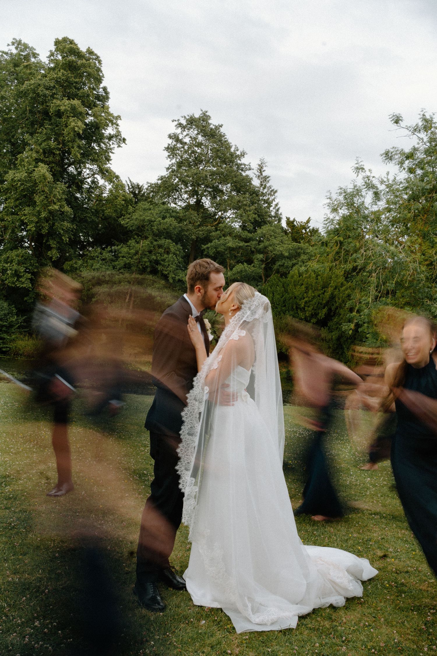 Long exposure photo of bride and groom kissing while wedding guests swirl around them in a lush garden setting.