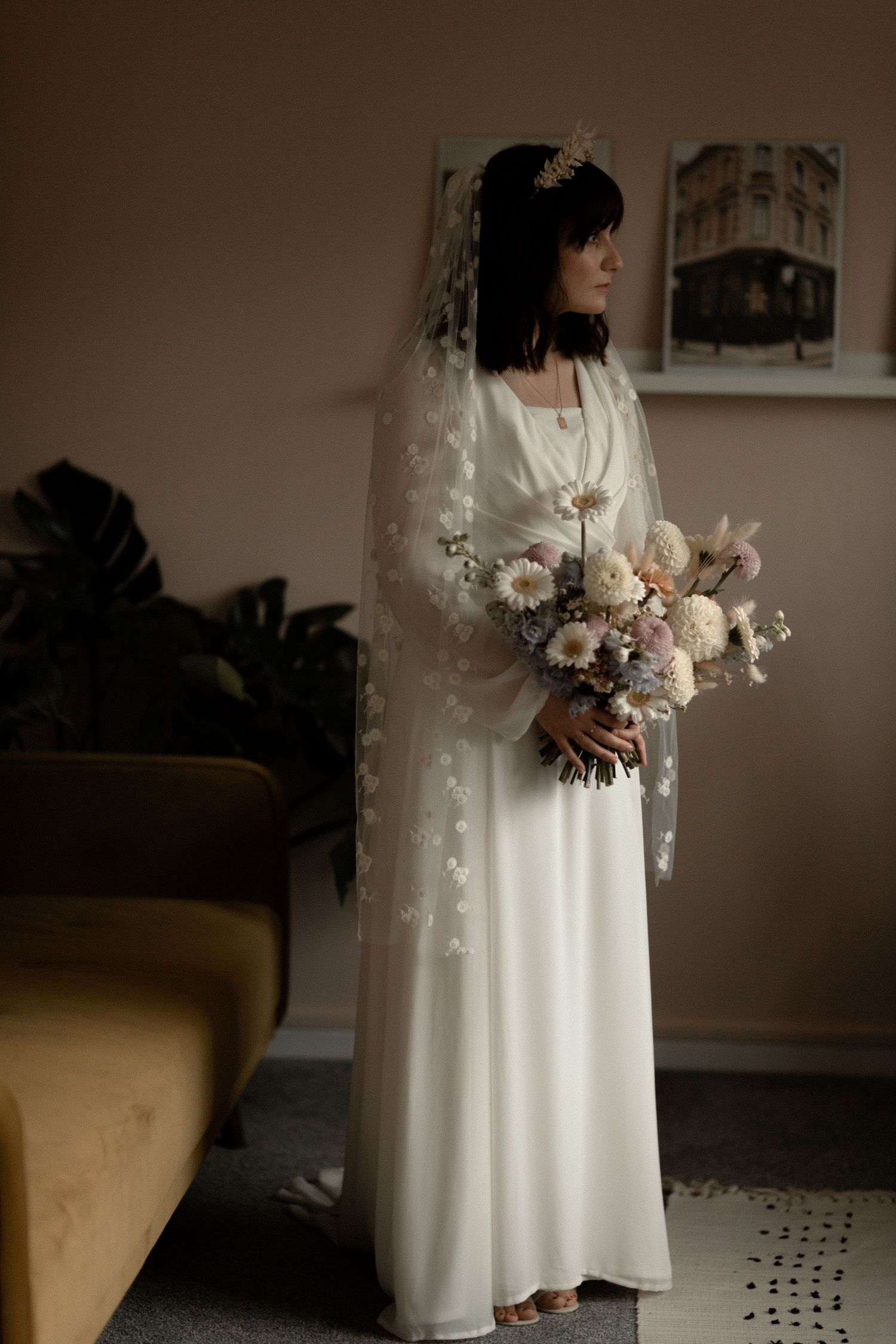 An elegant bride in a flowing white dress poses with a bouquet against a minimalist interior.
