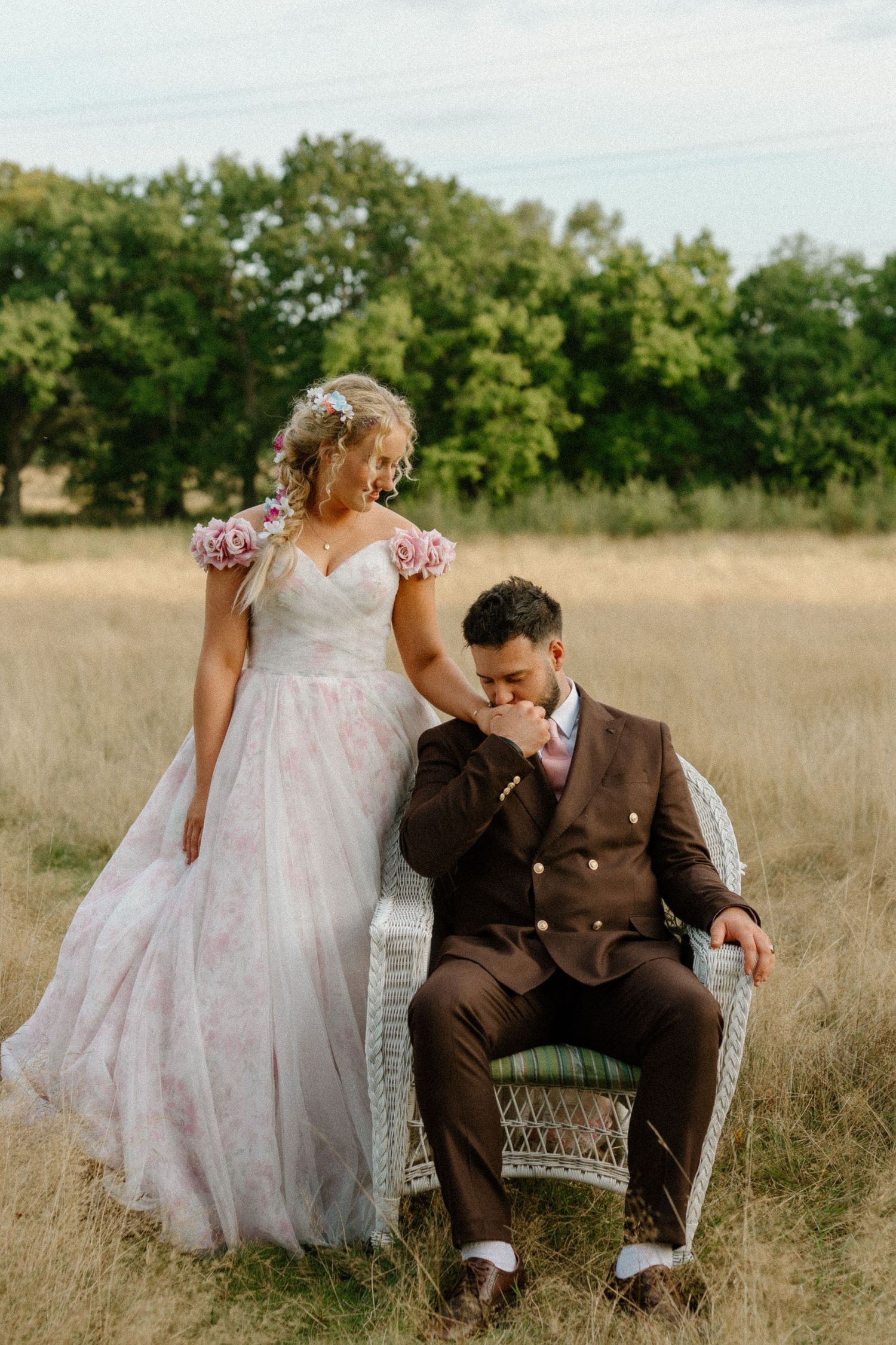 A romantic wedding photo in a golden field with a couple posing on vintage wicker furniture at sunset.