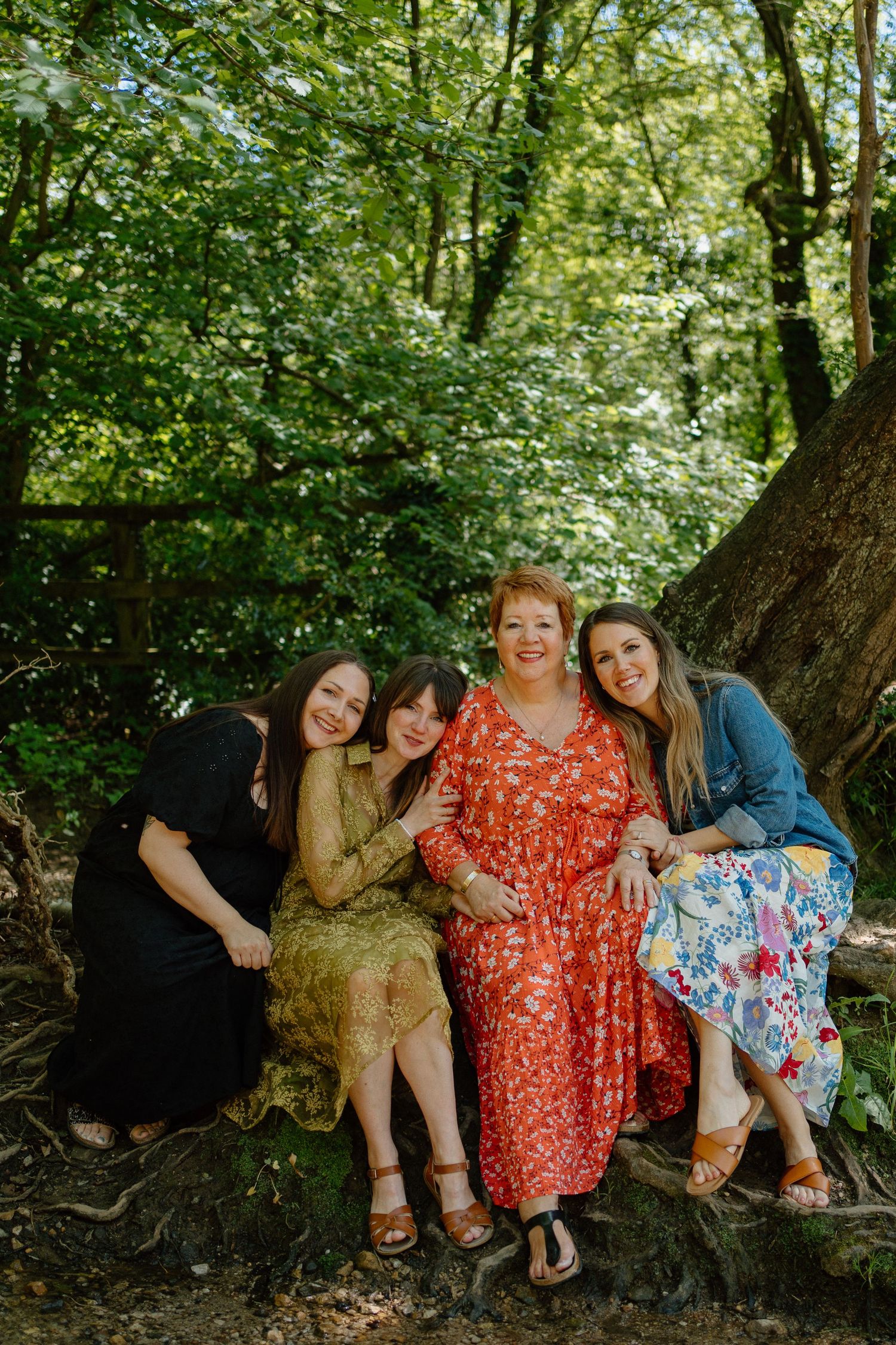 Four women smile together seated on tree roots in a lush green forest setting.