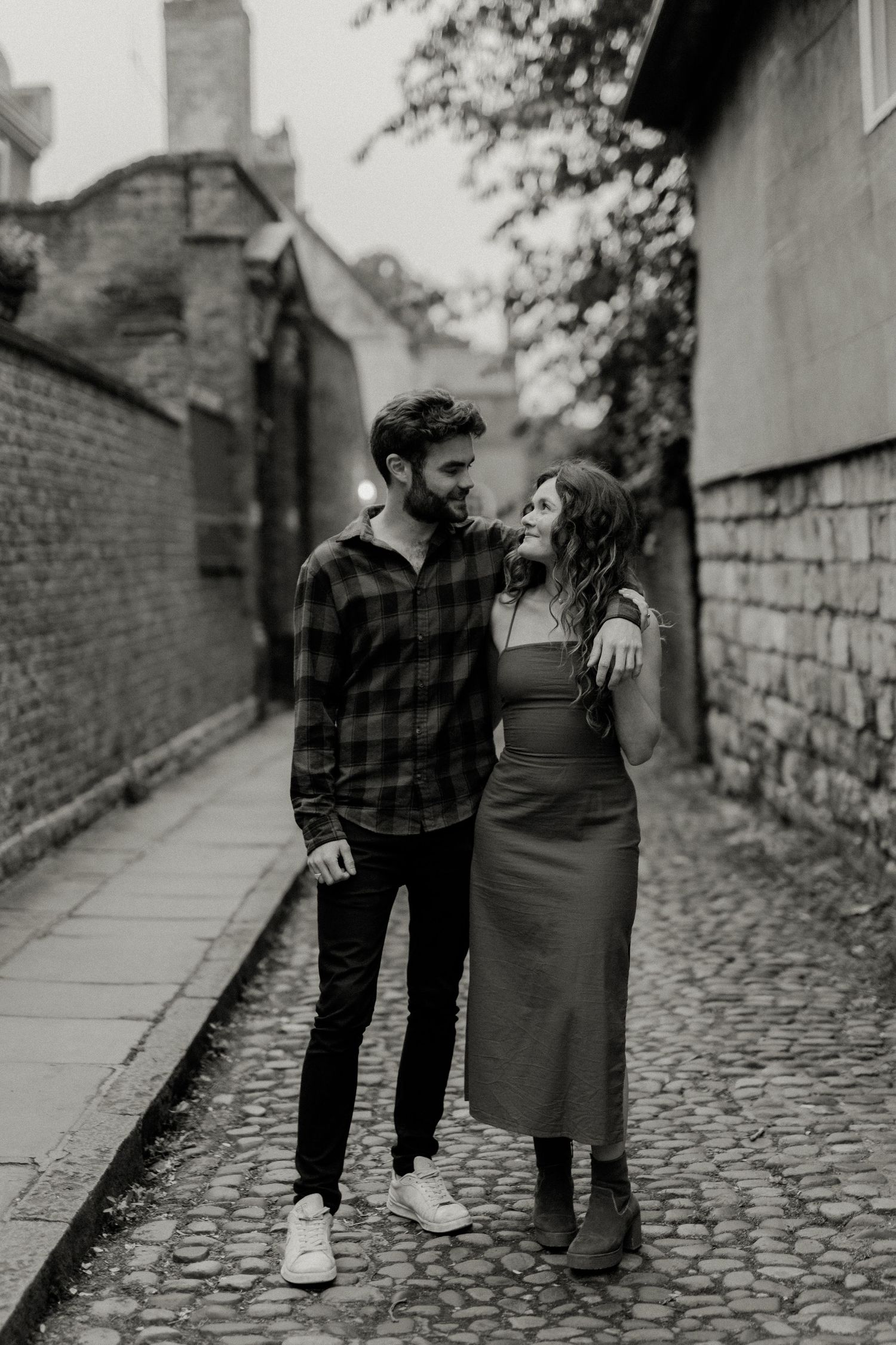 Black and white photo of a couple gazing at each other on a narrow cobblestone alleyway.