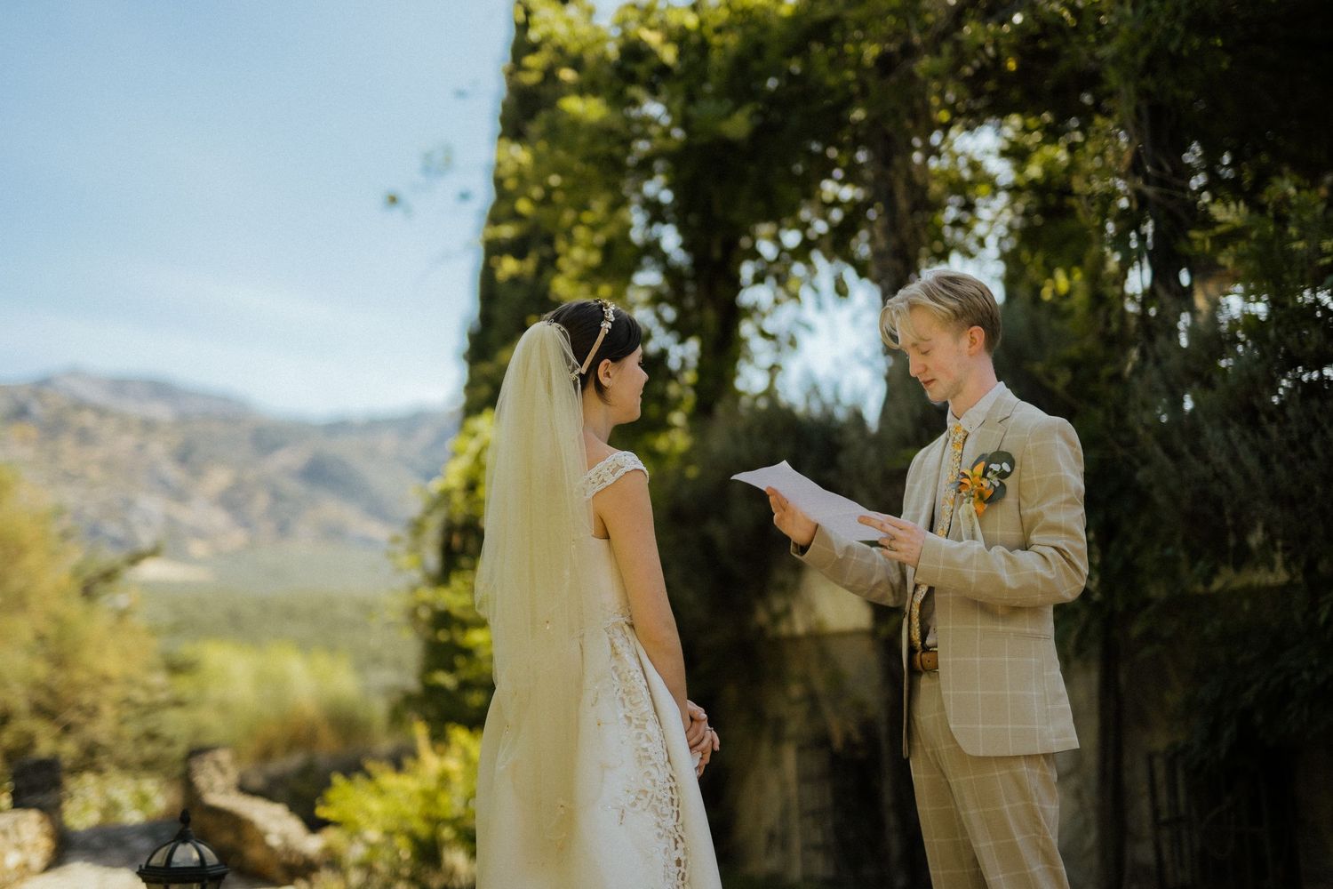 Groom reads wedding vows to bride in white dress and veil with mountain backdrop.