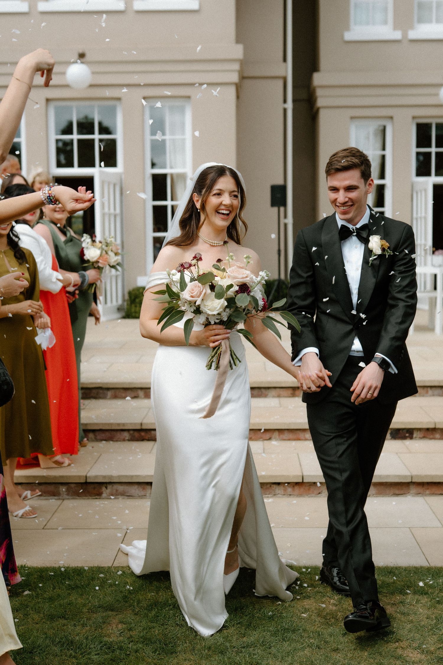 Joyful newlyweds walk through confetti shower outside elegant venue as guests celebrate their marriage.