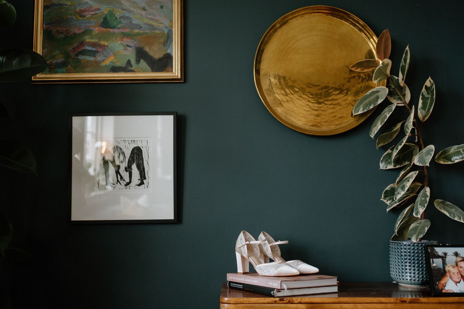 Wedding shoes and framed photo styled on a wooden dresser against a dark green wall with gold decor.