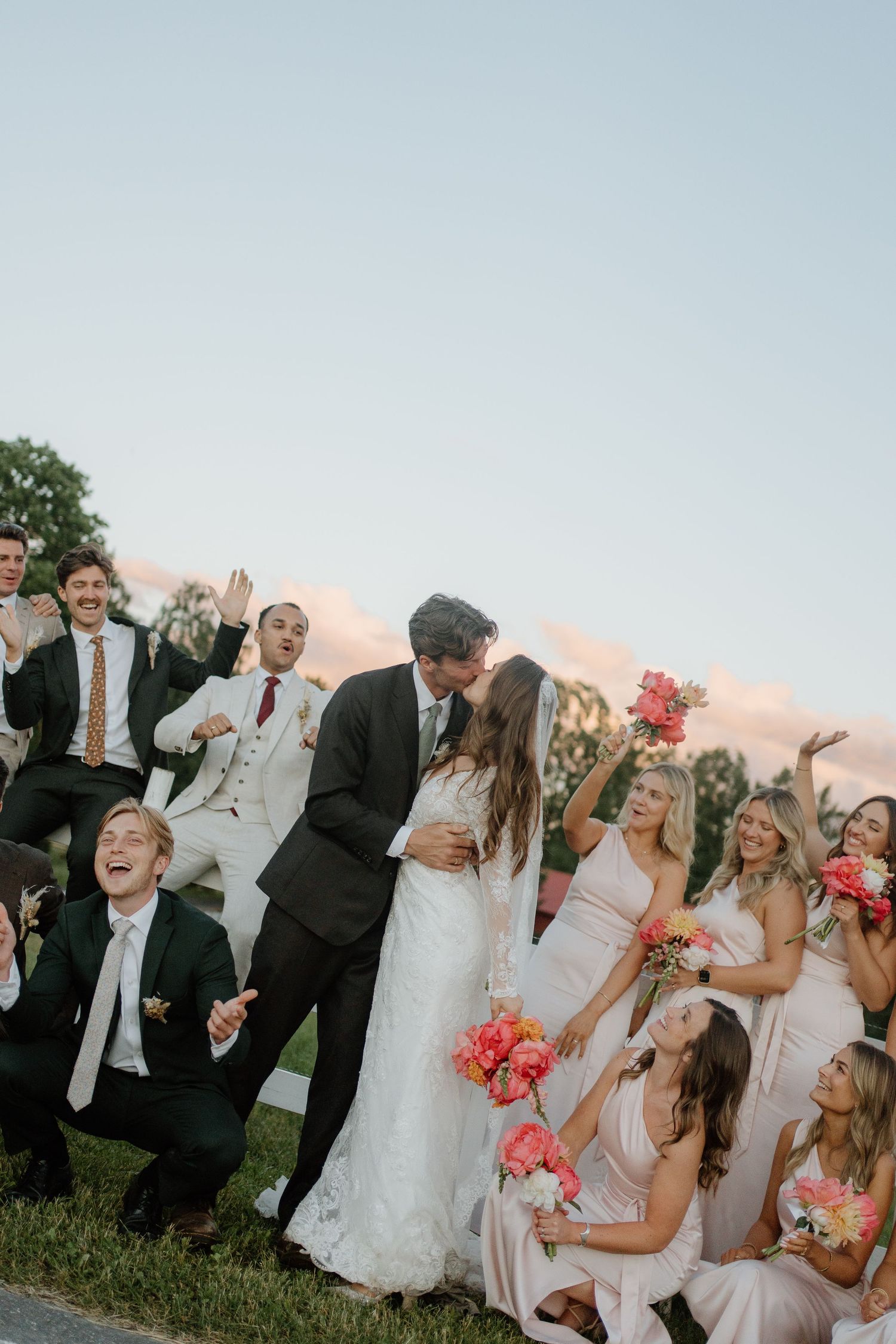 Wedding party celebrates as bride and groom share a kiss, bridesmaids holding coral bouquets in pink dresses.