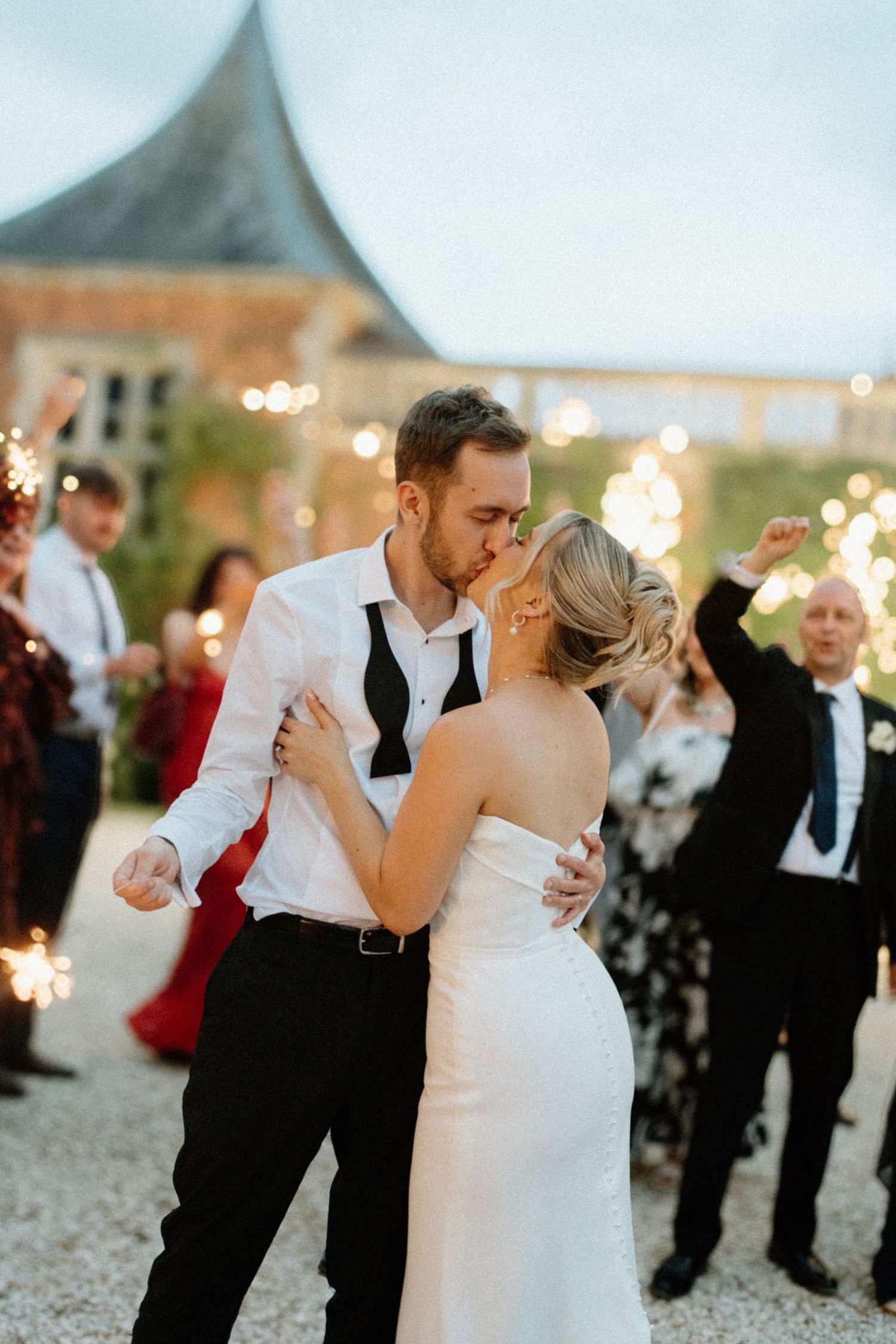 Bride and groom share passionate kiss surrounded by sparklers and celebrating guests outside elegant venue.