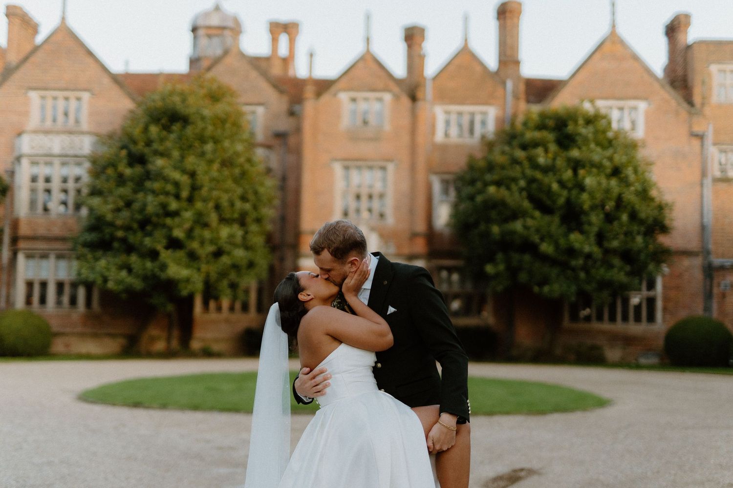 Newlyweds share romantic kiss in front of grand historic brick manor house at golden hour.