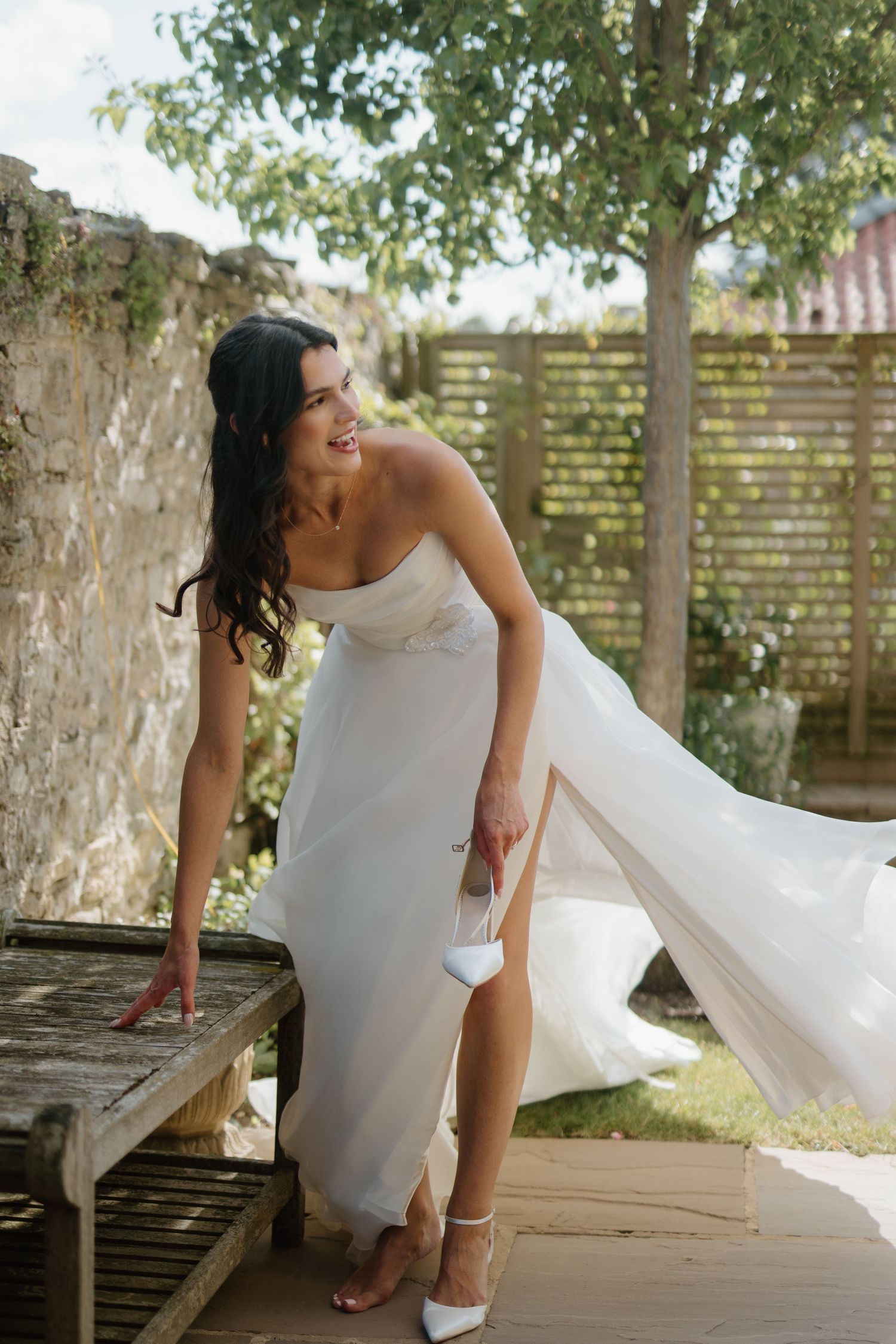 Radiant bride in strapless white gown with flowing train puts on heels in outdoor garden setting.