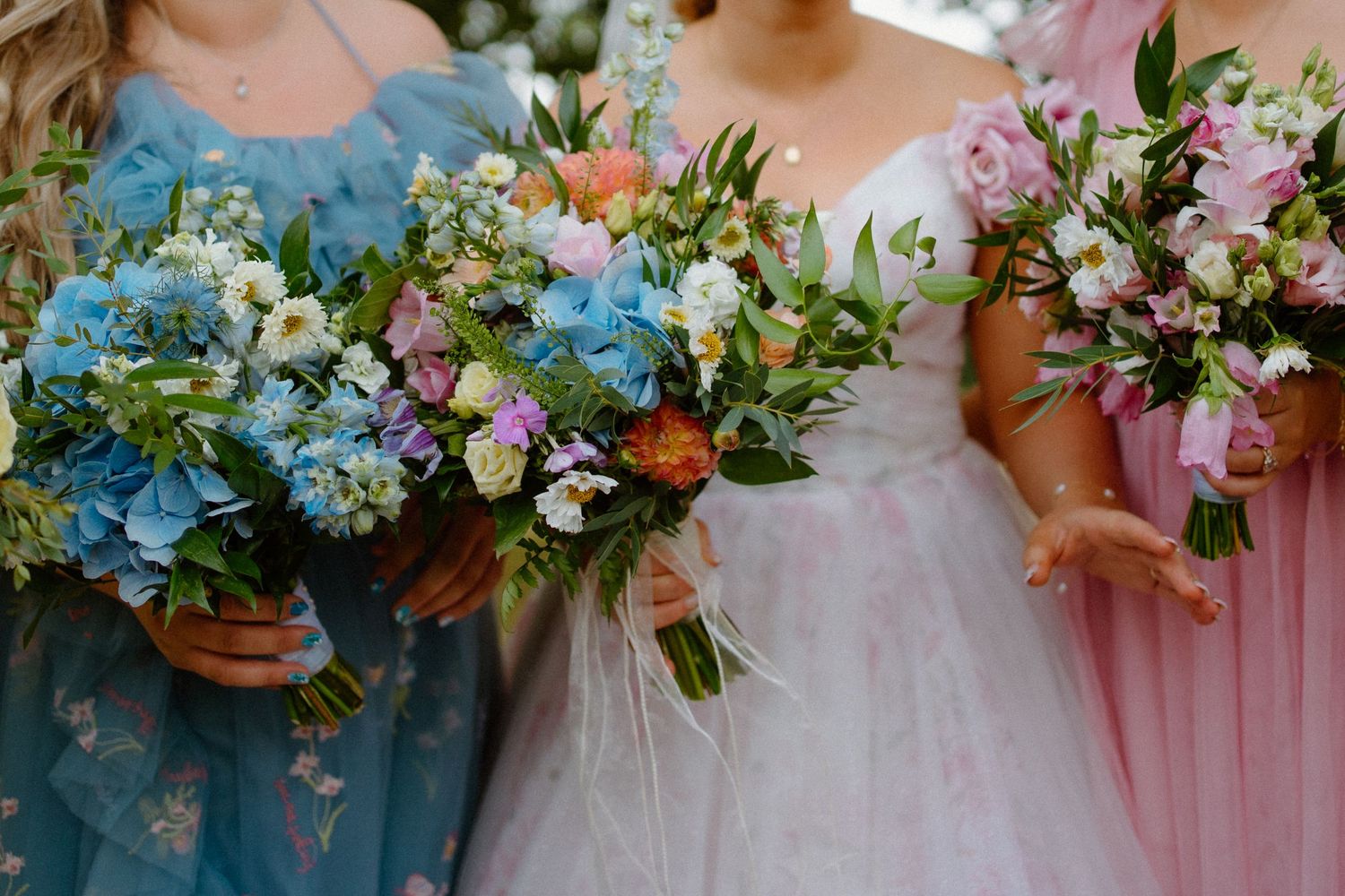 Bridesmaids in blue and pink gowns hold colorful wildflower bouquets at a wedding.