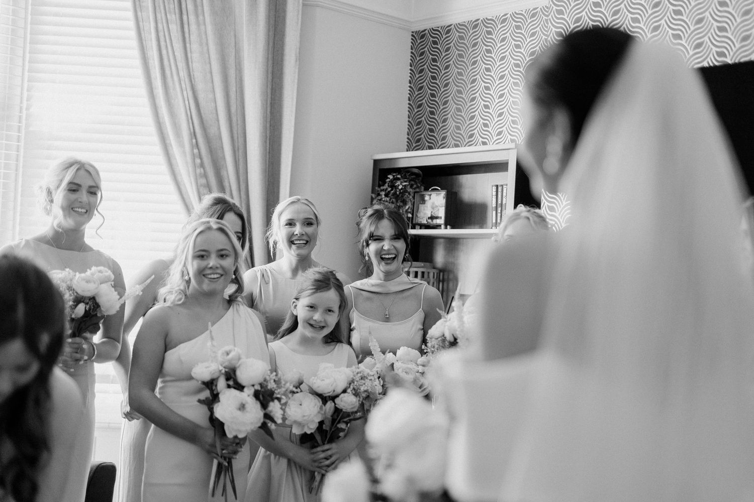 Black and white photo of bridesmaids reacting to bride in wedding dress during bridal preparations.