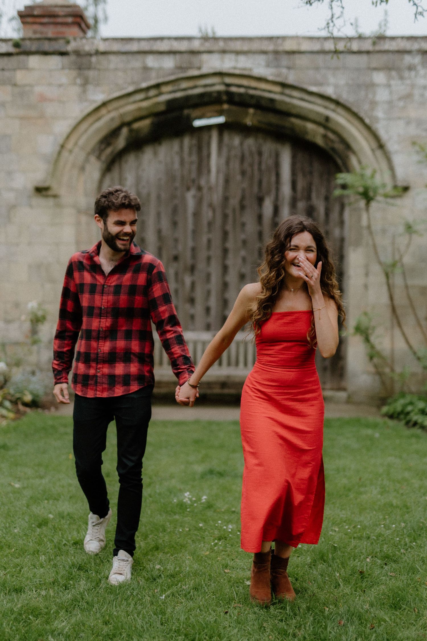 A couple holds hands while walking on grass in front of a rustic wooden door, wearing red dress and plaid shirt.