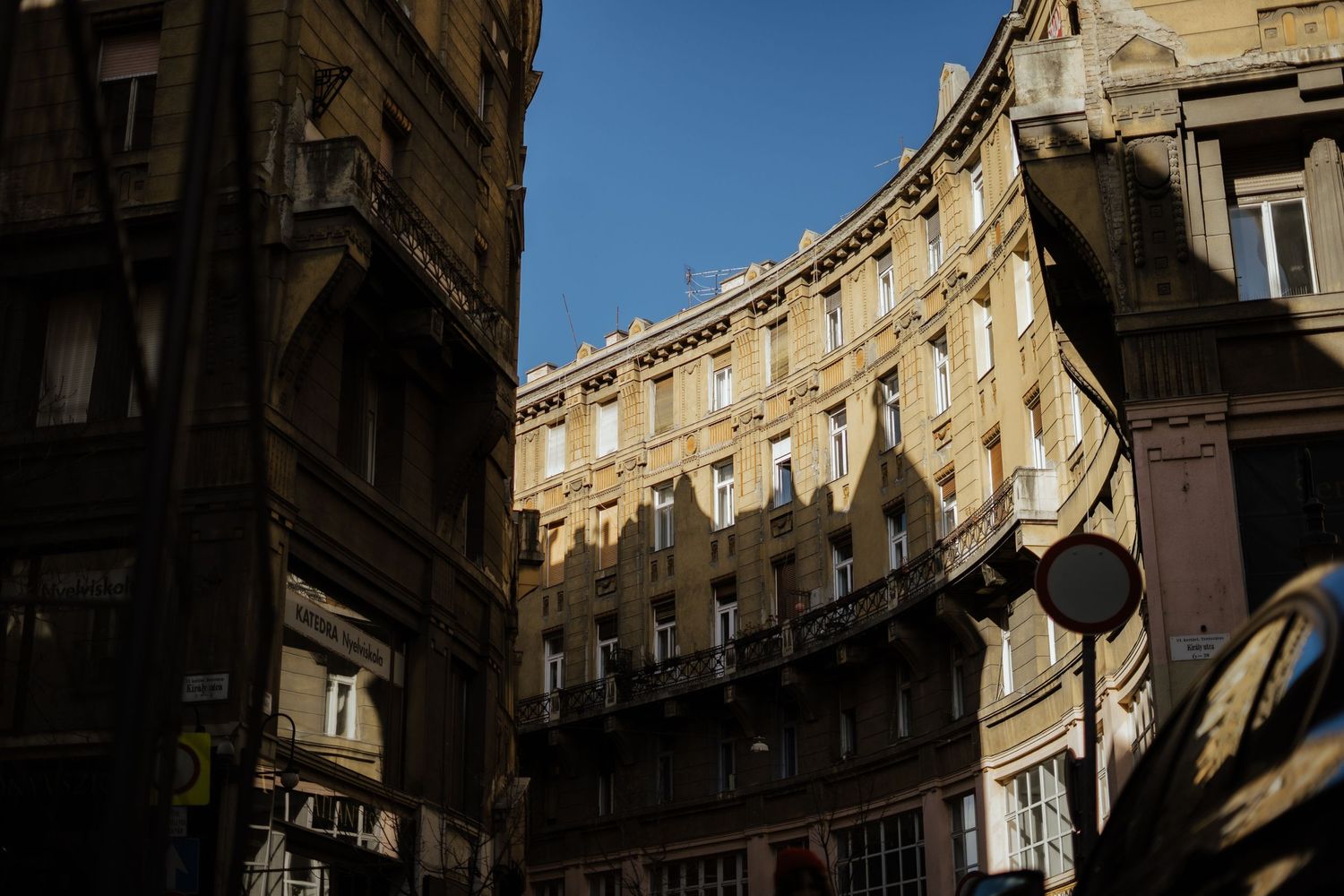 Curved historic building facade with ornate architecture and dramatic shadows cast across yellow walls in golden sunlight.
