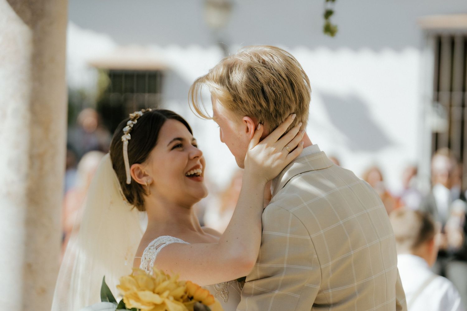 Laughing bride in veil holding yellow bouquet embraces blonde groom after wedding ceremony.