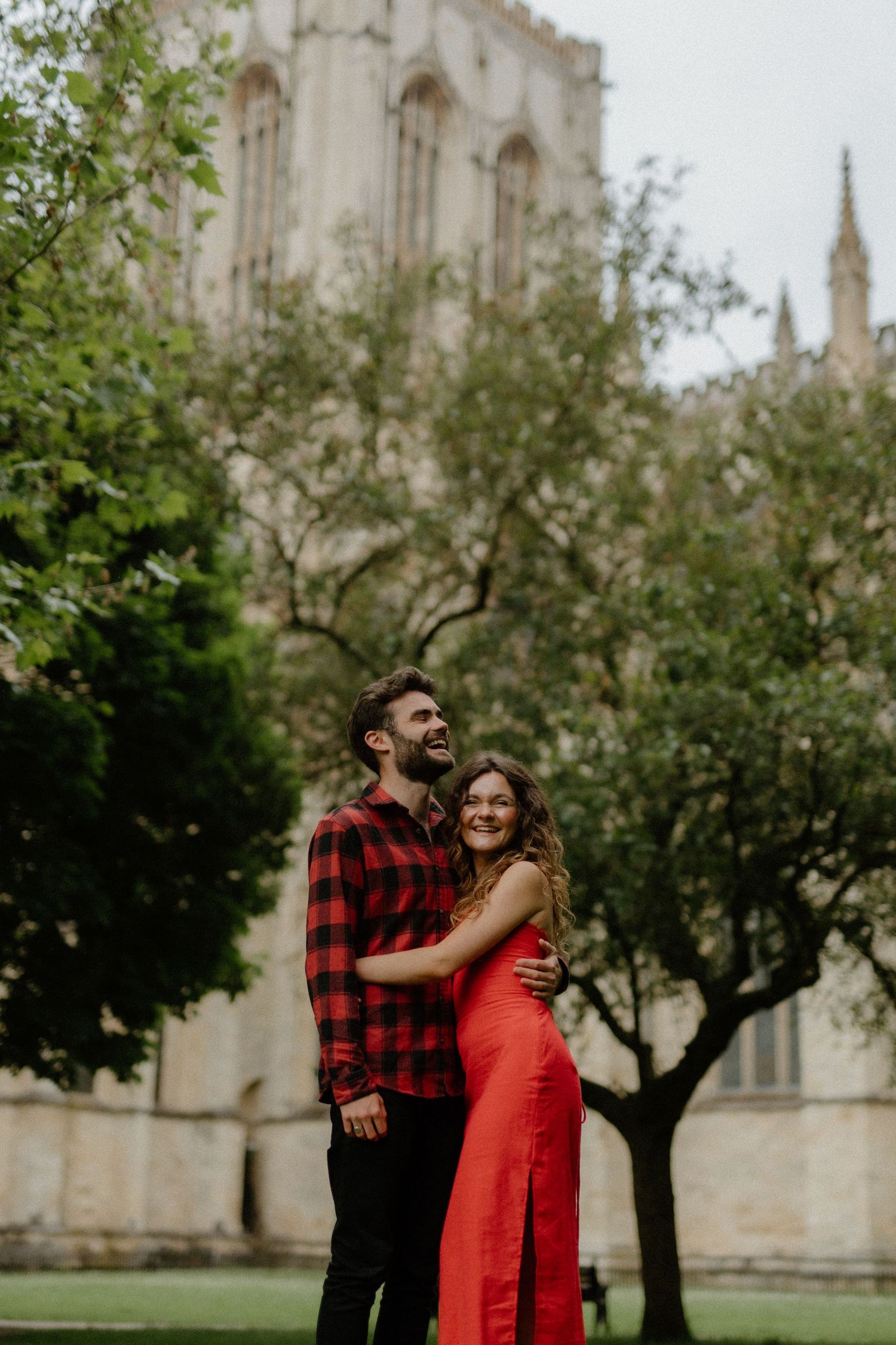 Happy couple embracing in a garden with a grand cathedral tower rising behind the trees.