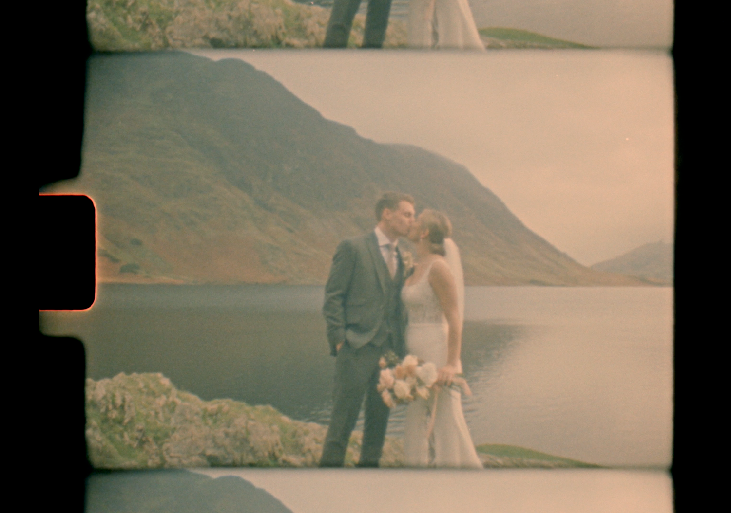 Vintage film-style photo of bride and groom kissing by a mountain lake with dramatic rocky landscape.