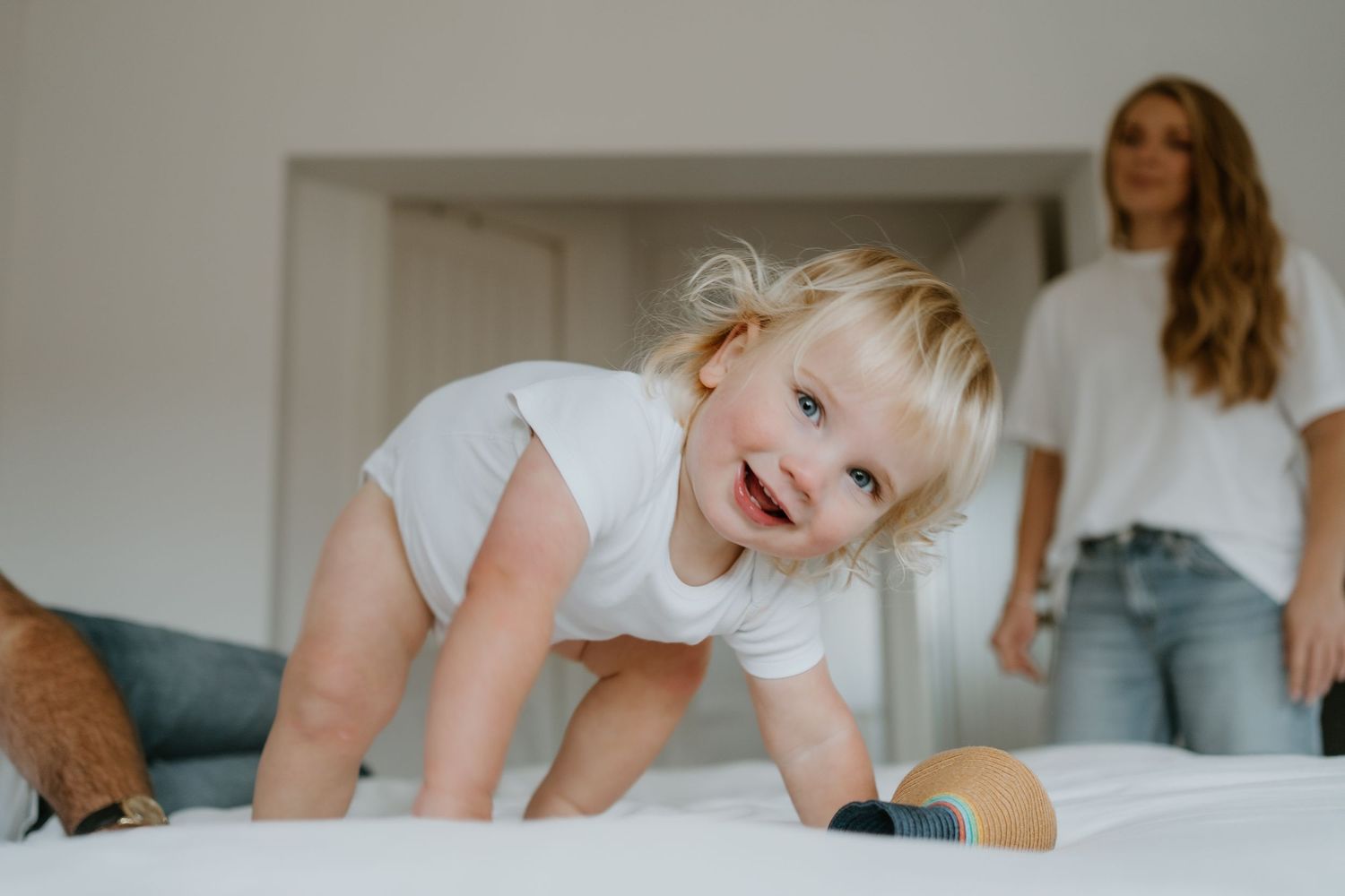 Happy blonde toddler crawling on white bed, smiling at camera with parents in background.