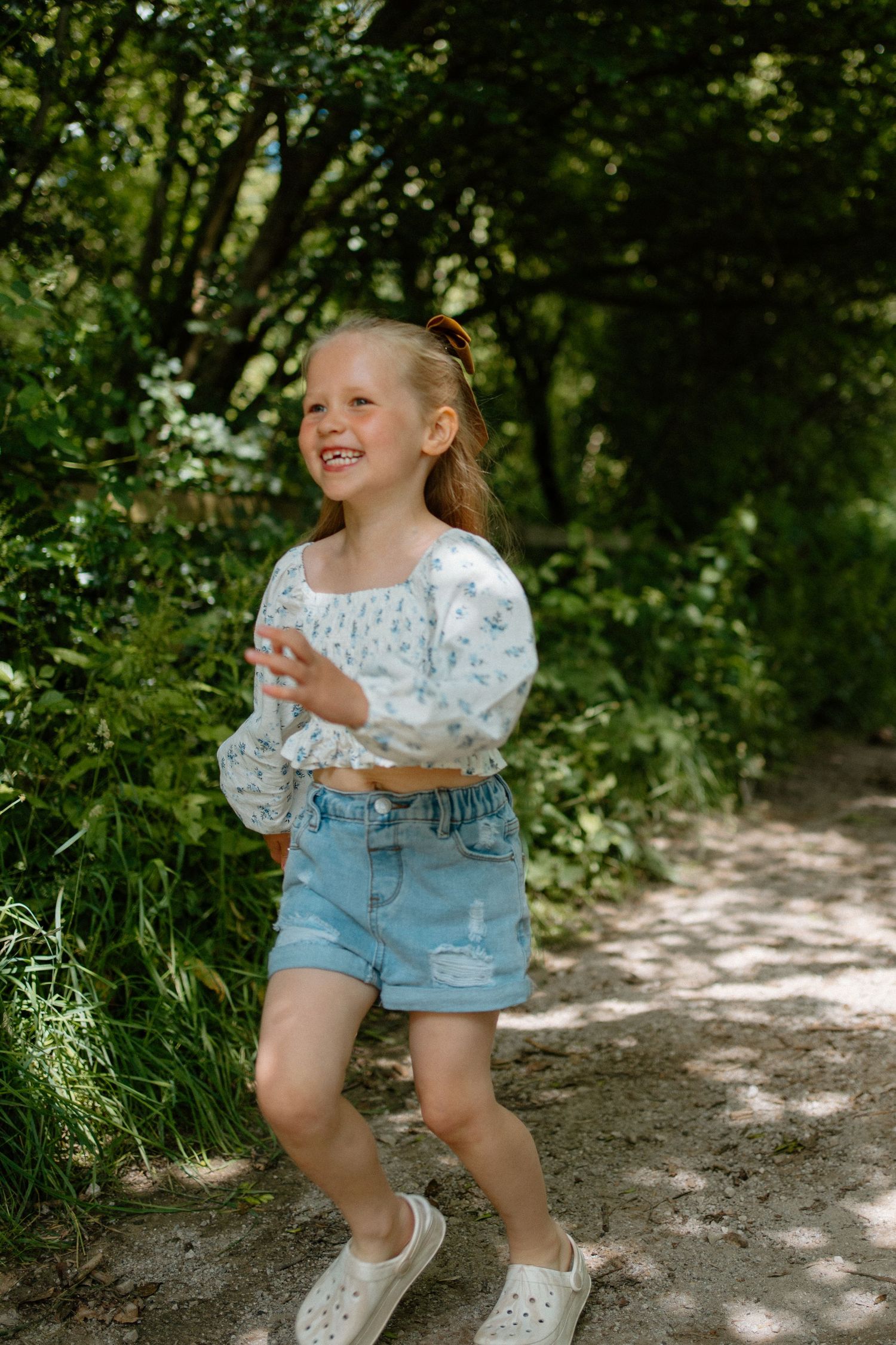 Happy young girl in floral top and denim shorts laughing while running along a lush green forest path.