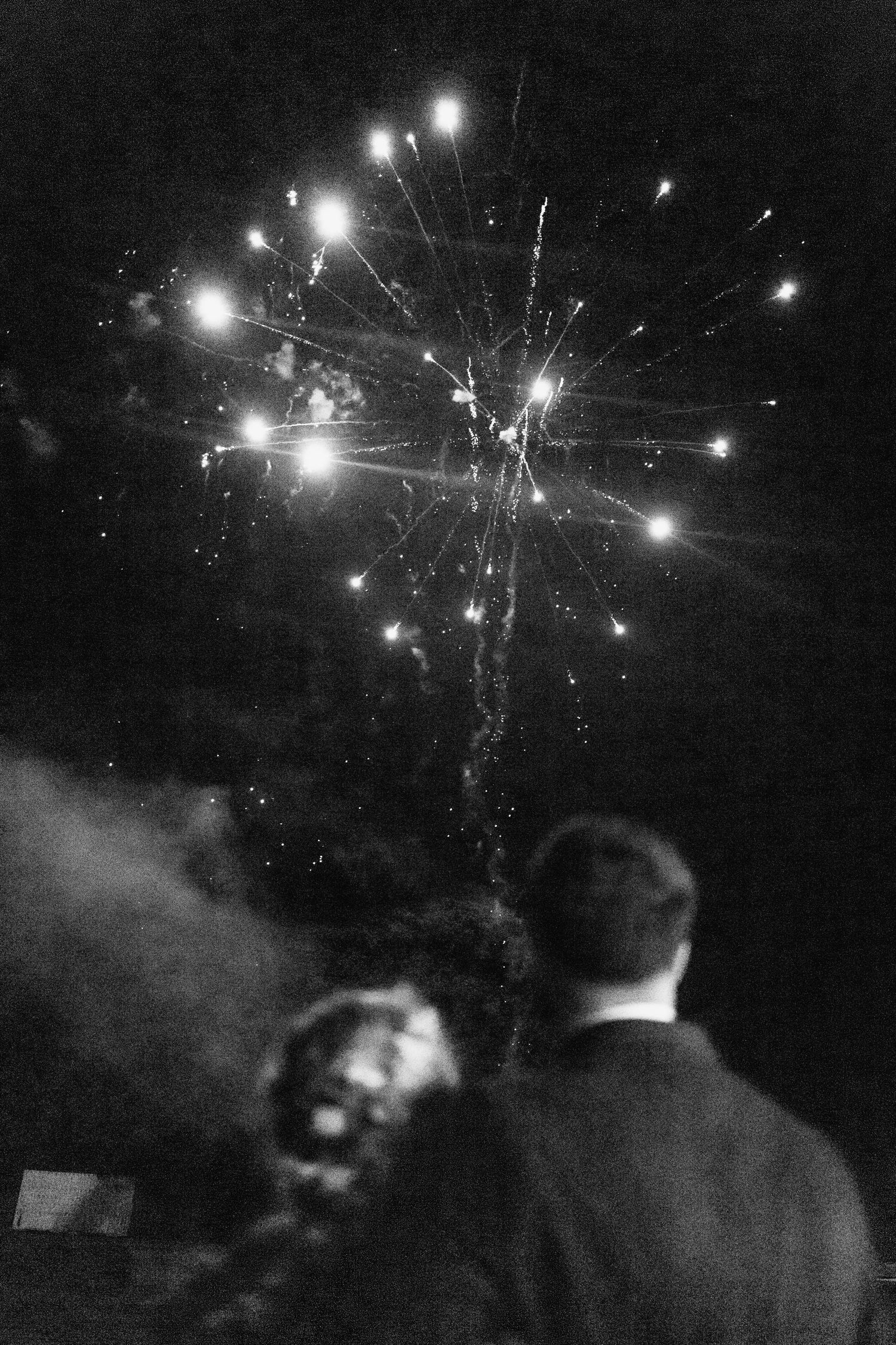 Black and white photo of newlyweds watching fireworks burst in the night sky at their wedding.