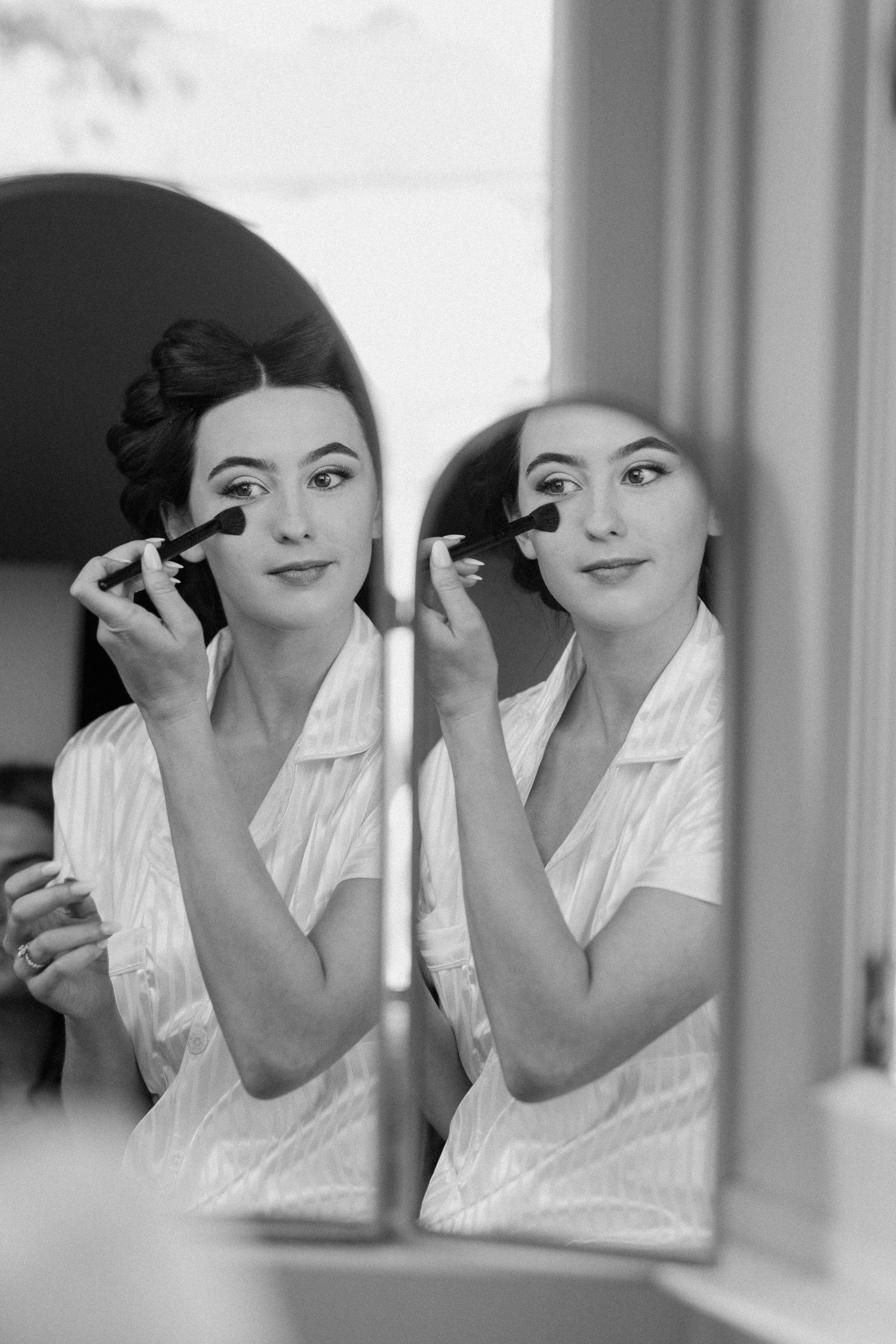 Black and white portrait of bride applying makeup brush while reflected in mirror before wedding.