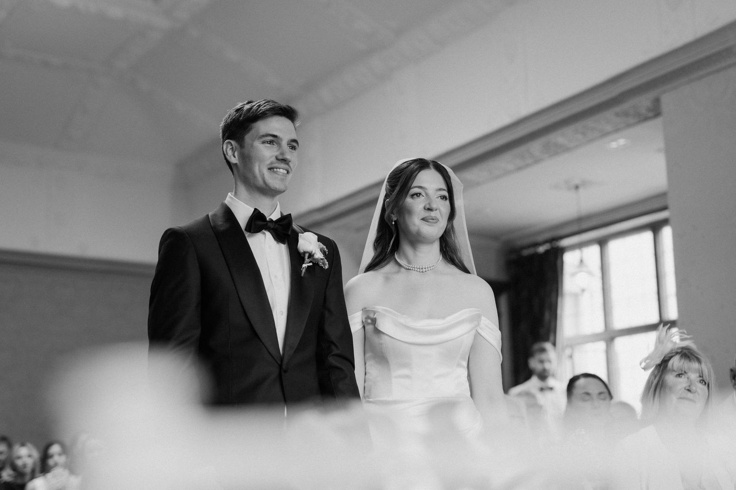 Black and white photo of bride and groom standing together smiling during elegant wedding ceremony.