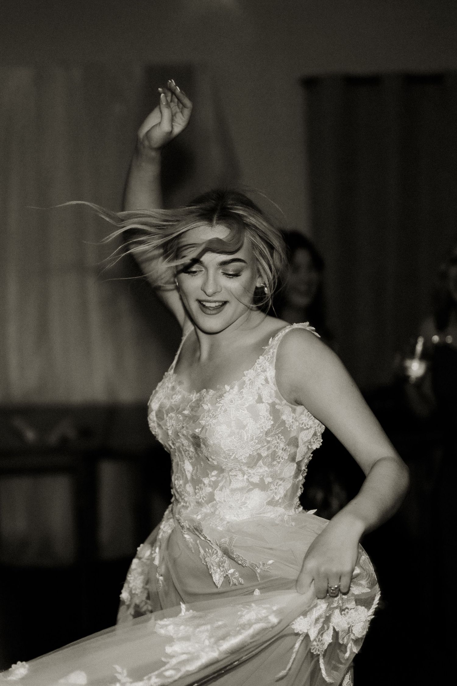Black and white photo of bride in lace wedding gown dancing joyfully with hair flowing at reception.