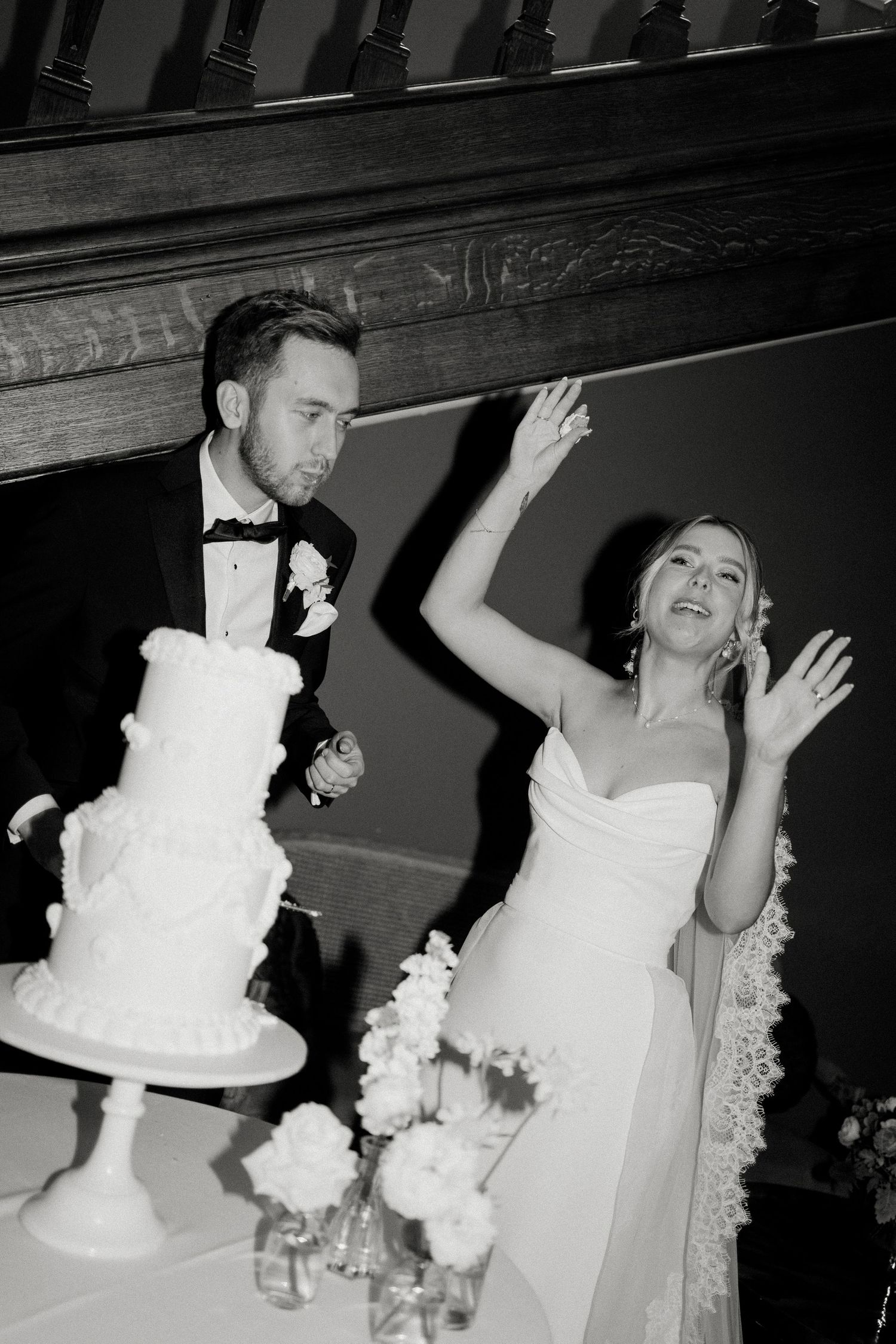 Black and white photo of bride and groom cutting their elegant tiered white wedding cake during evening reception.