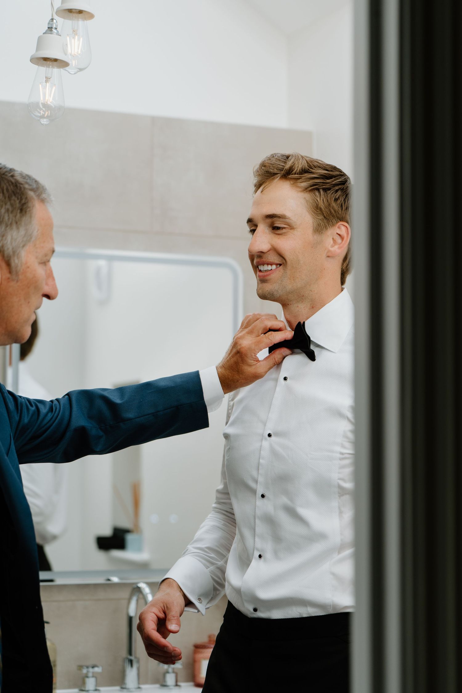 Older man helps smiling groom adjust his black bow tie before the wedding ceremony.