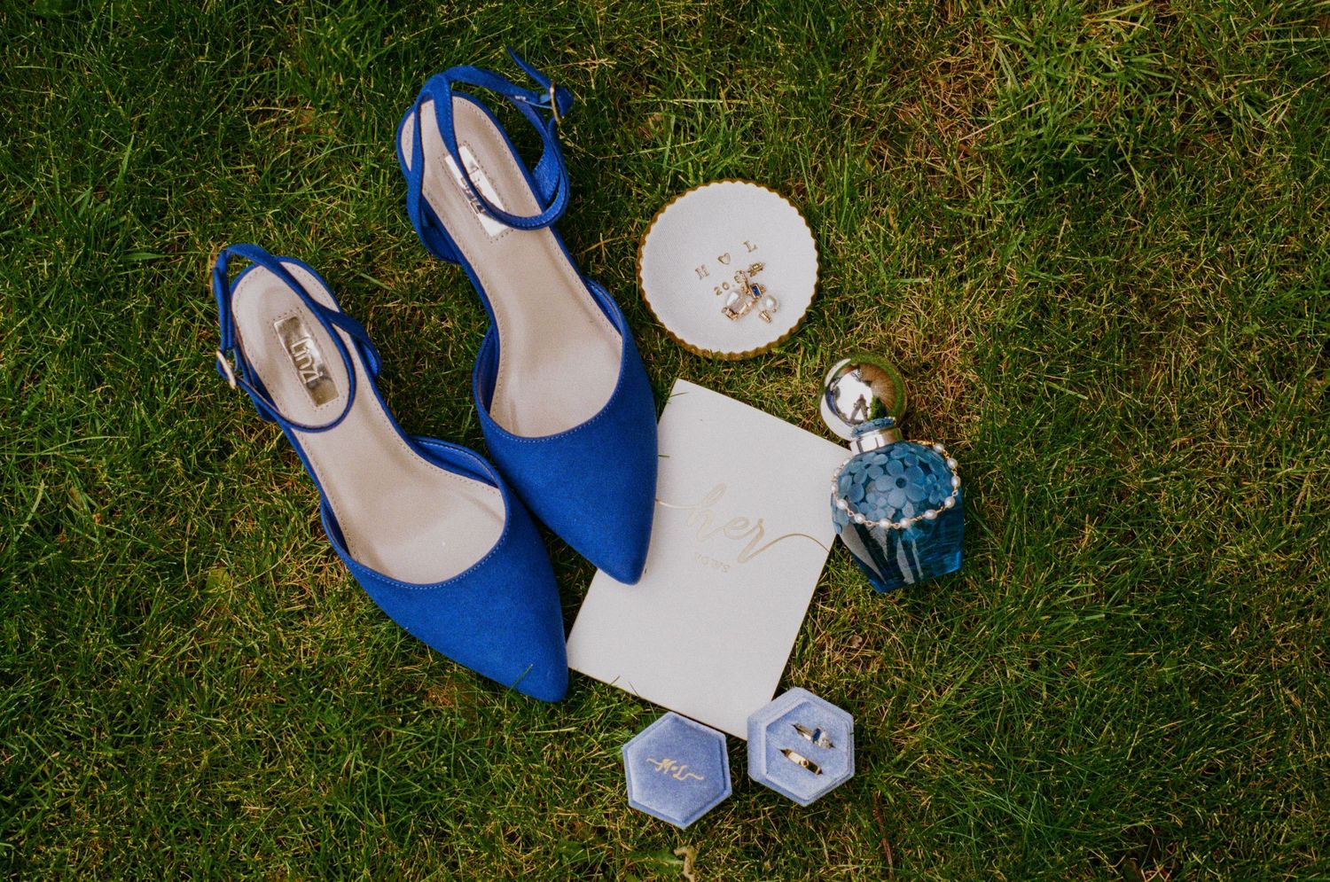 Blue suede wedding heels beside invitation, ring boxes, and perfume bottle on green grass.