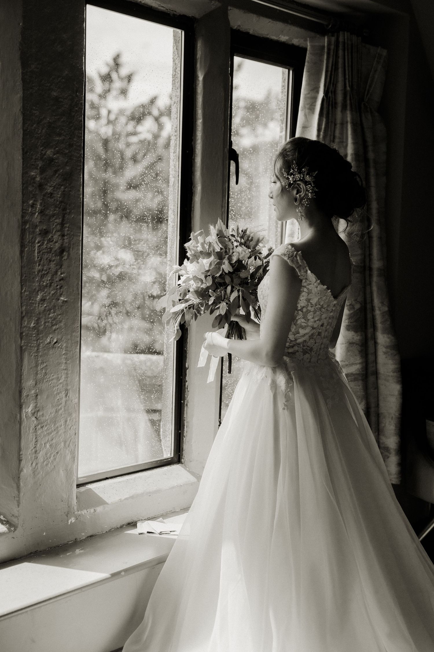 Bride in white gown holding bouquet gazes out window in black and white wedding photo.