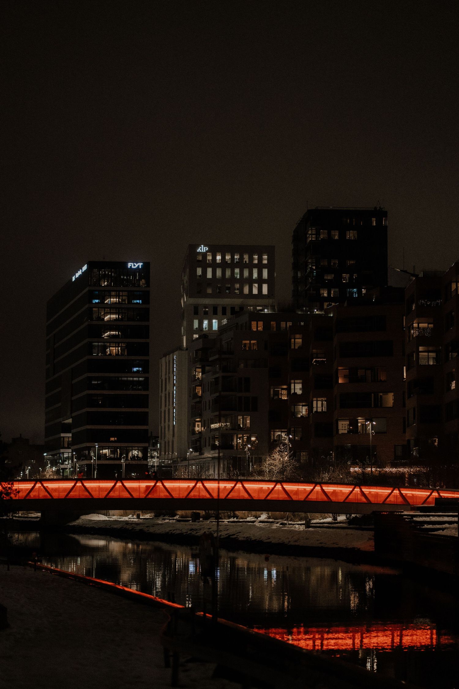 Red-lit bridge reflects in calm water at night with illuminated city buildings rising in the background.