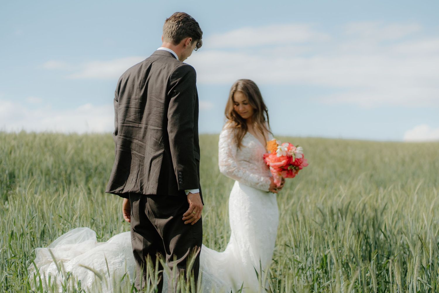 Bride in long-sleeve lace gown holds colorful bouquet with groom in green wheat field.