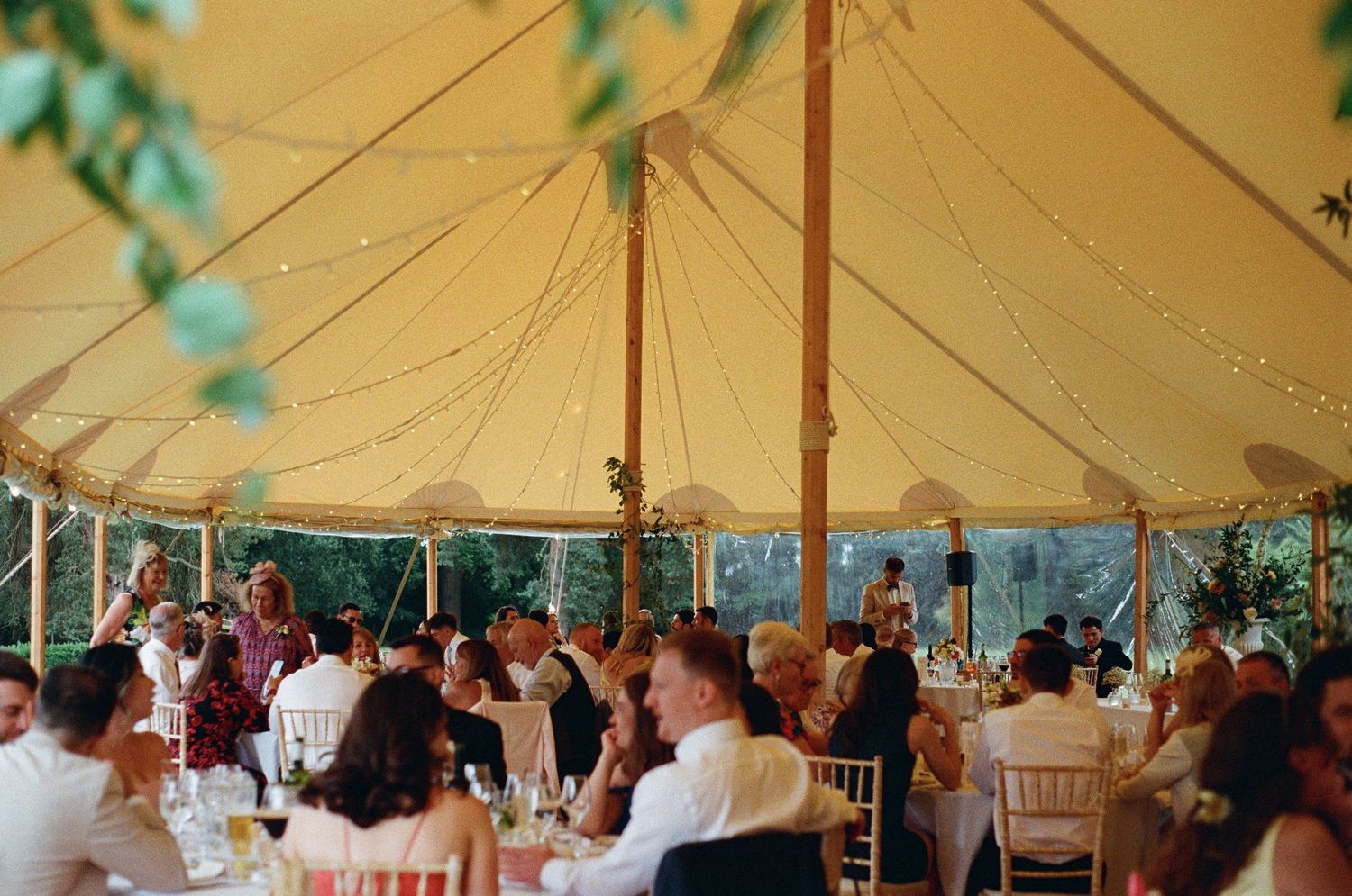 Wedding guests seated at round tables under a warm sailcloth tent with fairy lights at reception.