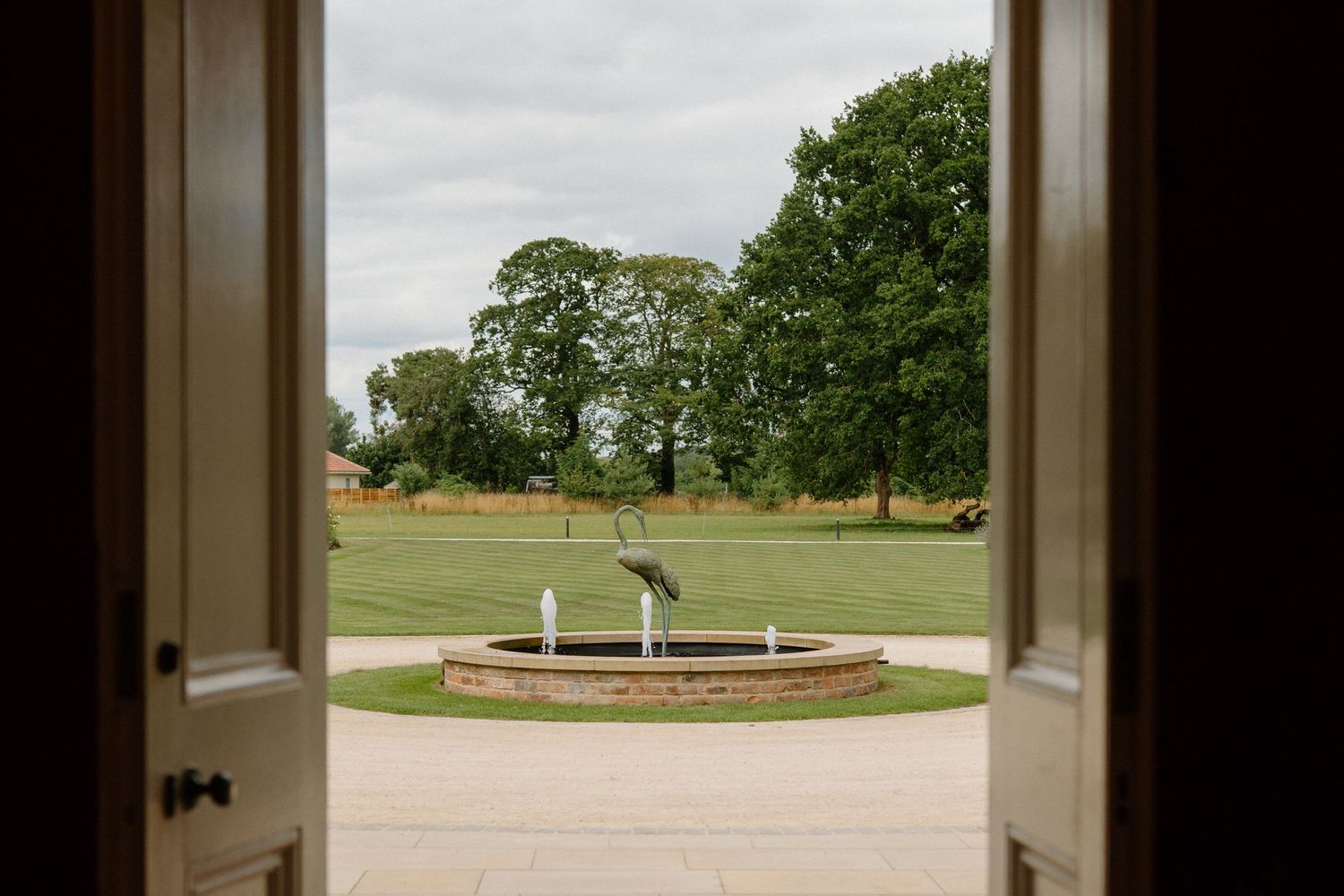 View through open wooden doors of elegant estate garden with circular fountain and lush green trees.
