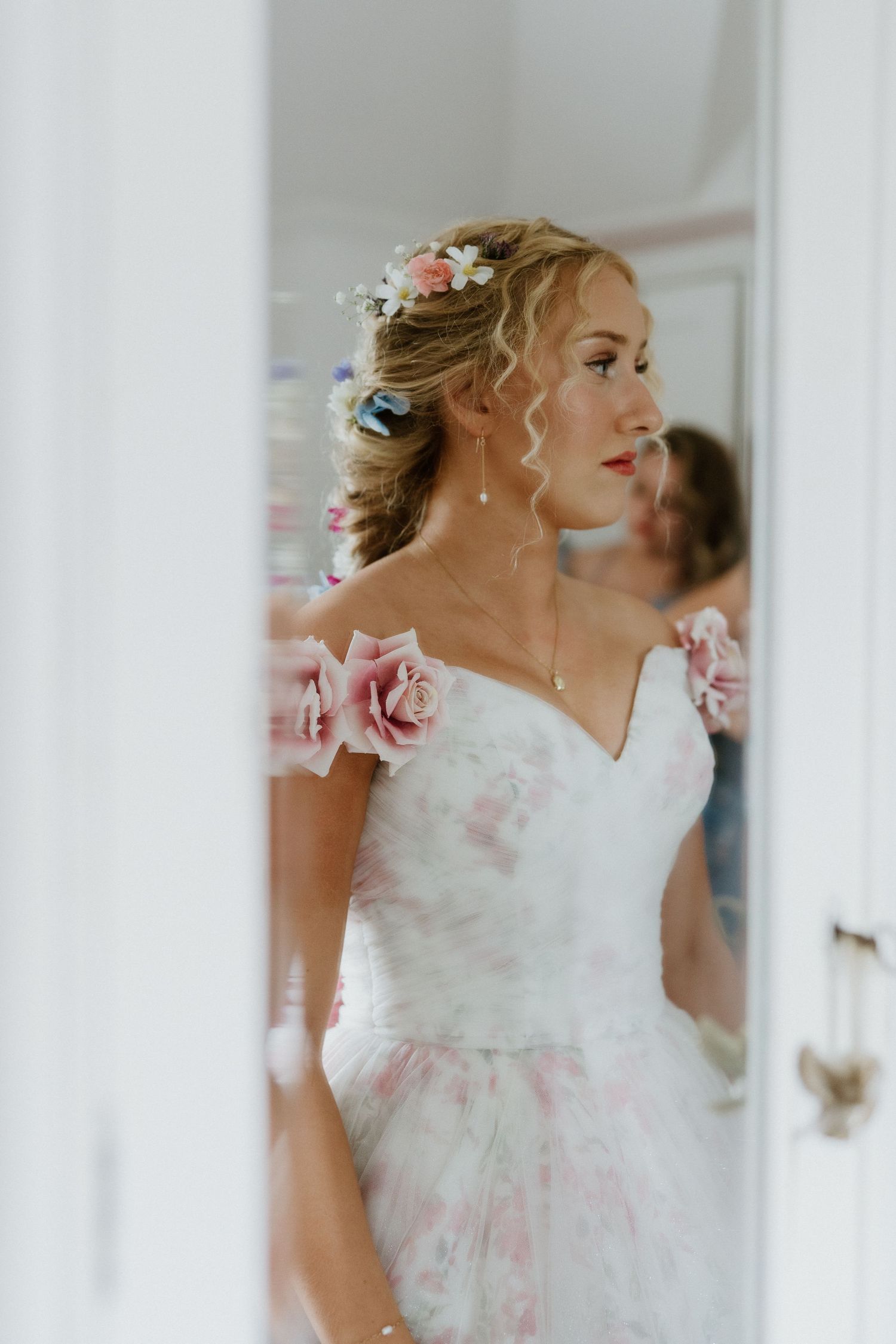 Elegant bride in pink floral gown with rose shoulder detail reflected in a mirror before wedding.