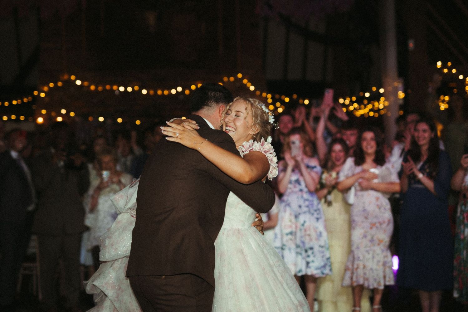 Bride and groom share a romantic embrace on the dance floor as wedding guests clap and celebrate.