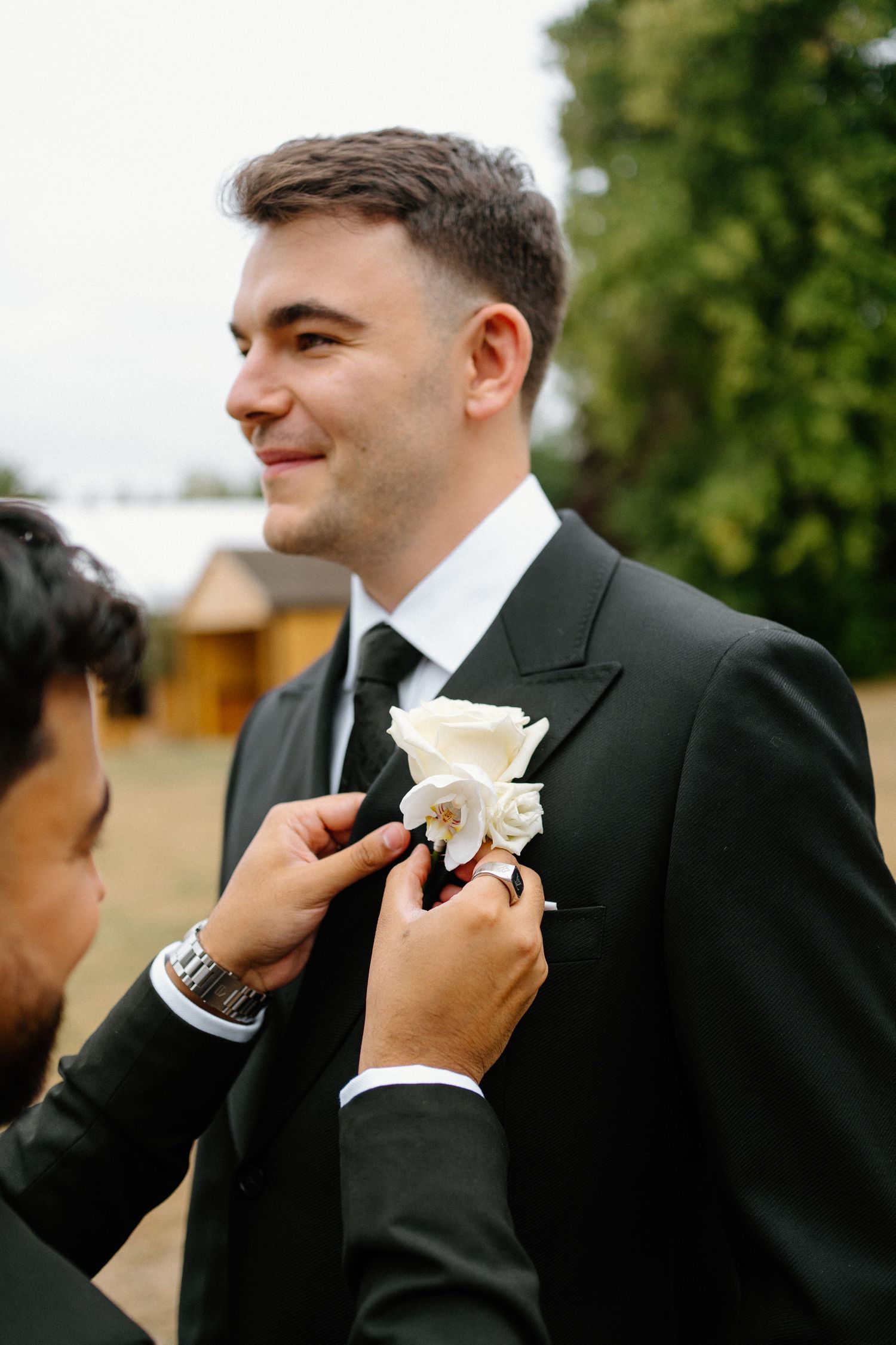 Groomsman pinning a white rose boutonniere onto the groom's black suit lapel outdoors.