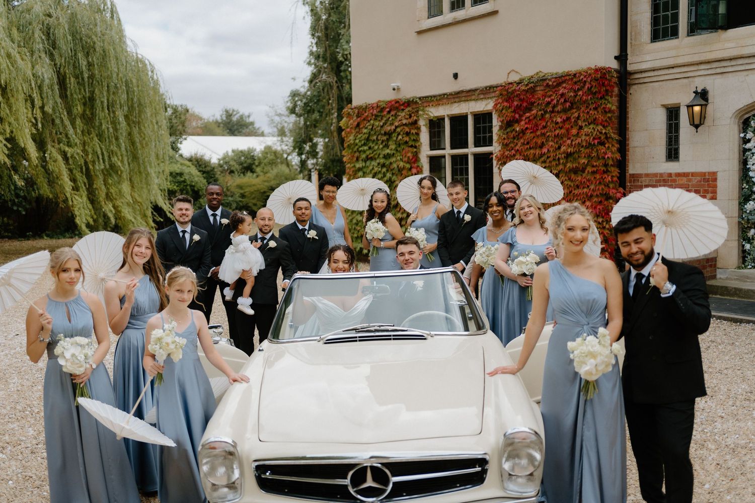 Wedding party posing with a classic white Mercedes convertible, bridesmaids in blue dresses holding white parasols.