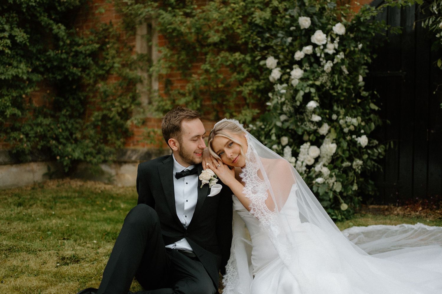 Bride and groom sitting together on grass in front of a beautiful white floral arch at their wedding venue.