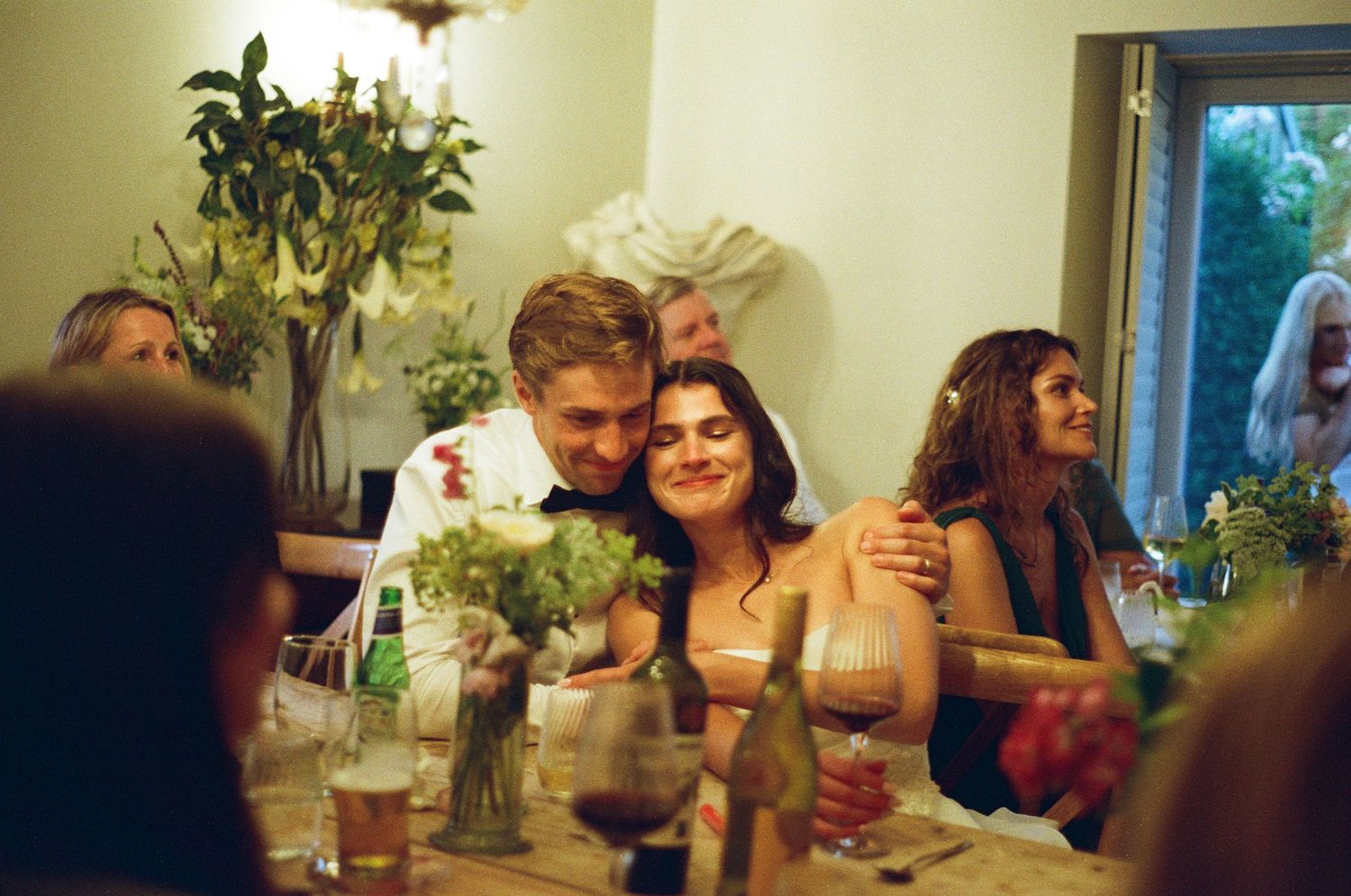 Wedding guests laugh and embrace at a candlelit reception dinner table with floral centerpieces.