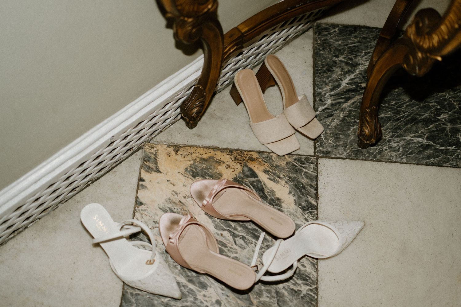 Two pairs of elegant bridal heels arranged on a marble floor beside ornate furniture.