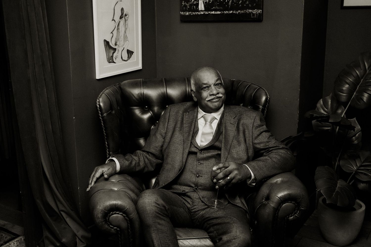 A man in a tweed suit relaxes in a leather armchair in a moody, dimly lit room with jazz artwork.