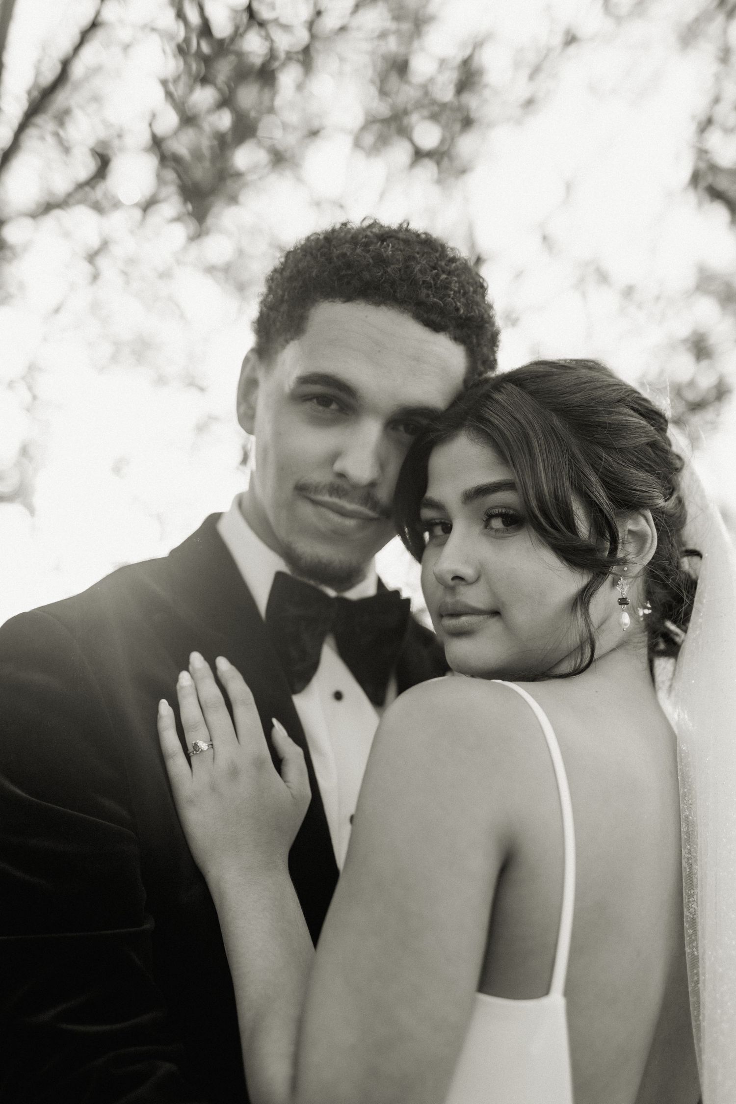 Black and white portrait of bride and groom embracing under trees on their wedding day.