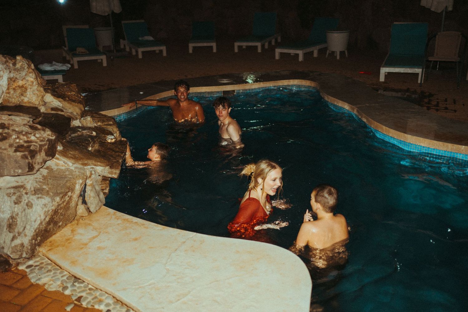 Group of wedding guests enjoying a nighttime swim in an outdoor pool surrounded by rock features.