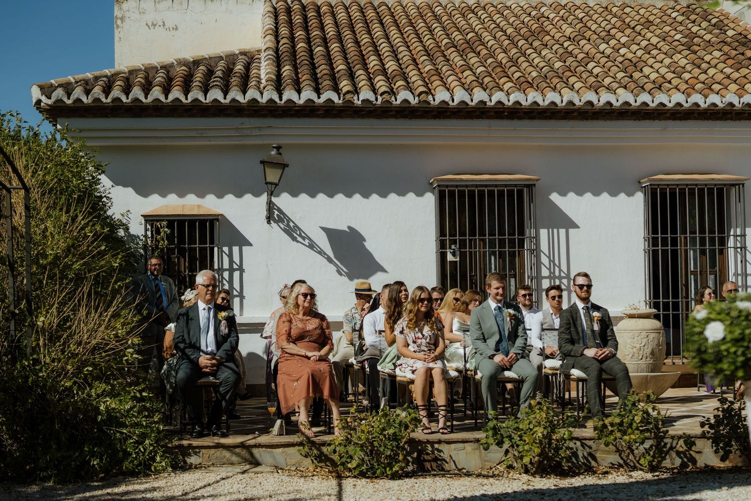 Wedding guests seated on benches outside a whitewashed Spanish villa during a sunny outdoor ceremony.