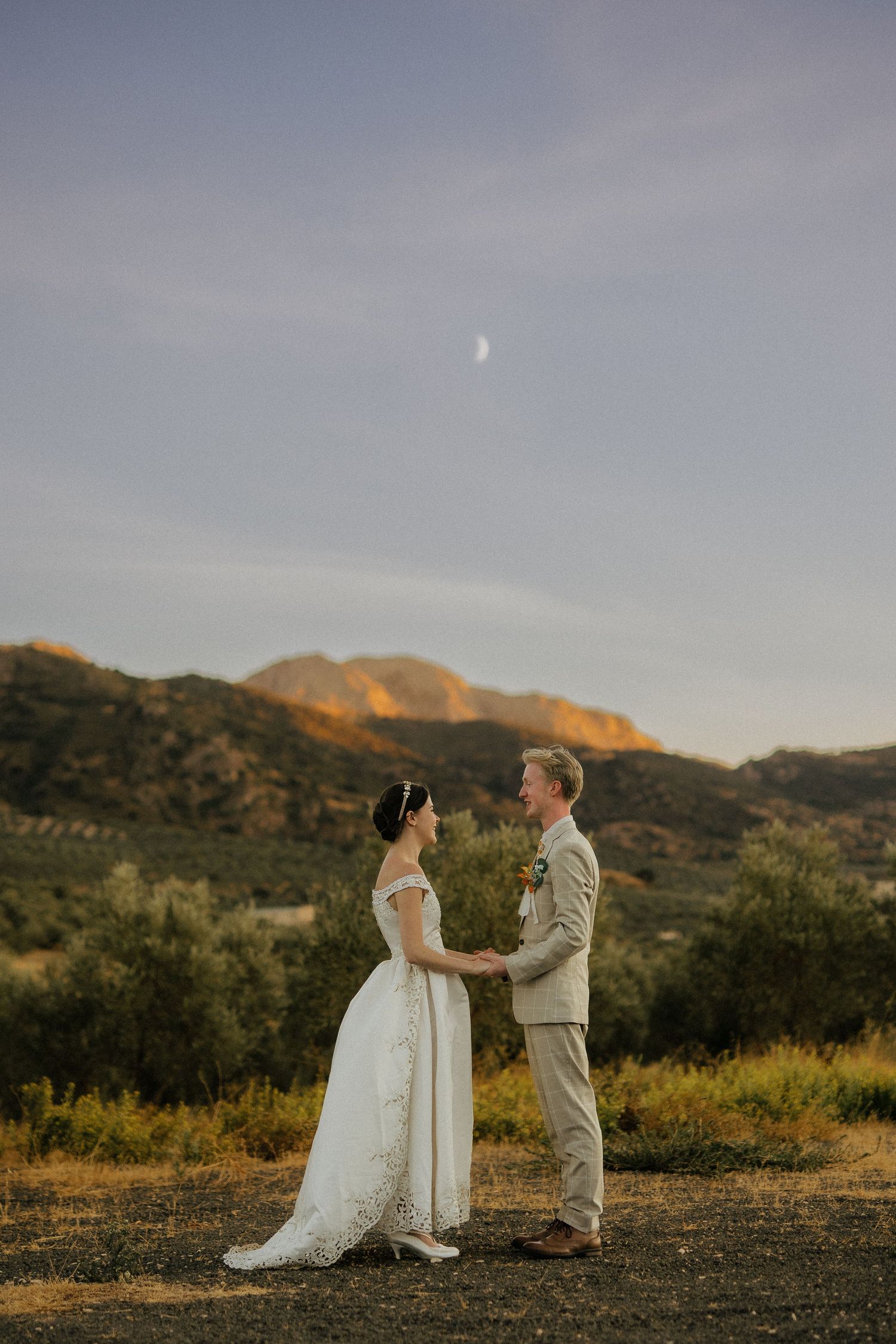 Bride and groom hold hands at sunset with golden mountain landscape and crescent moon above them.