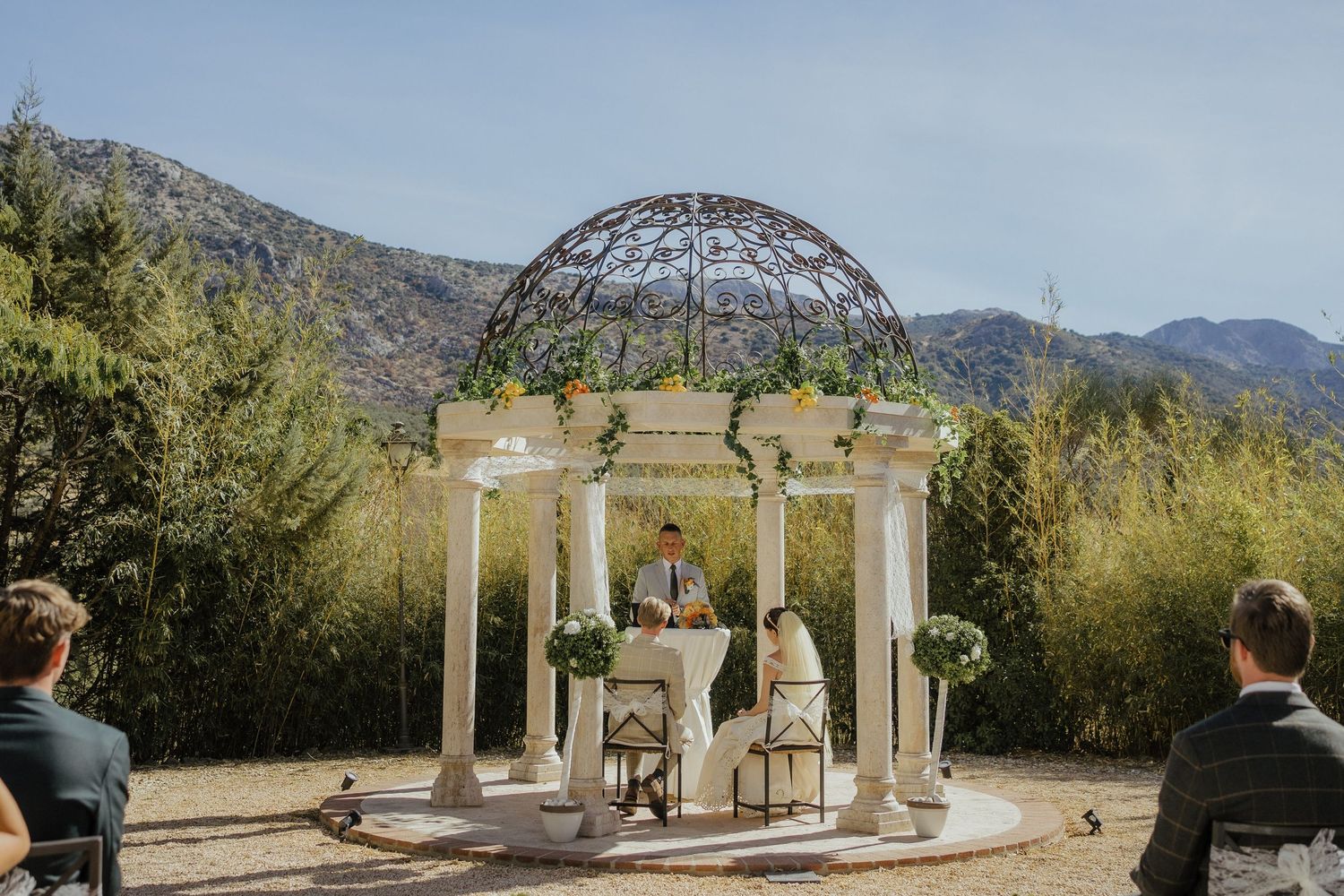 Elegant outdoor wedding ceremony under a domed gazebo with mountain backdrop and lush greenery.