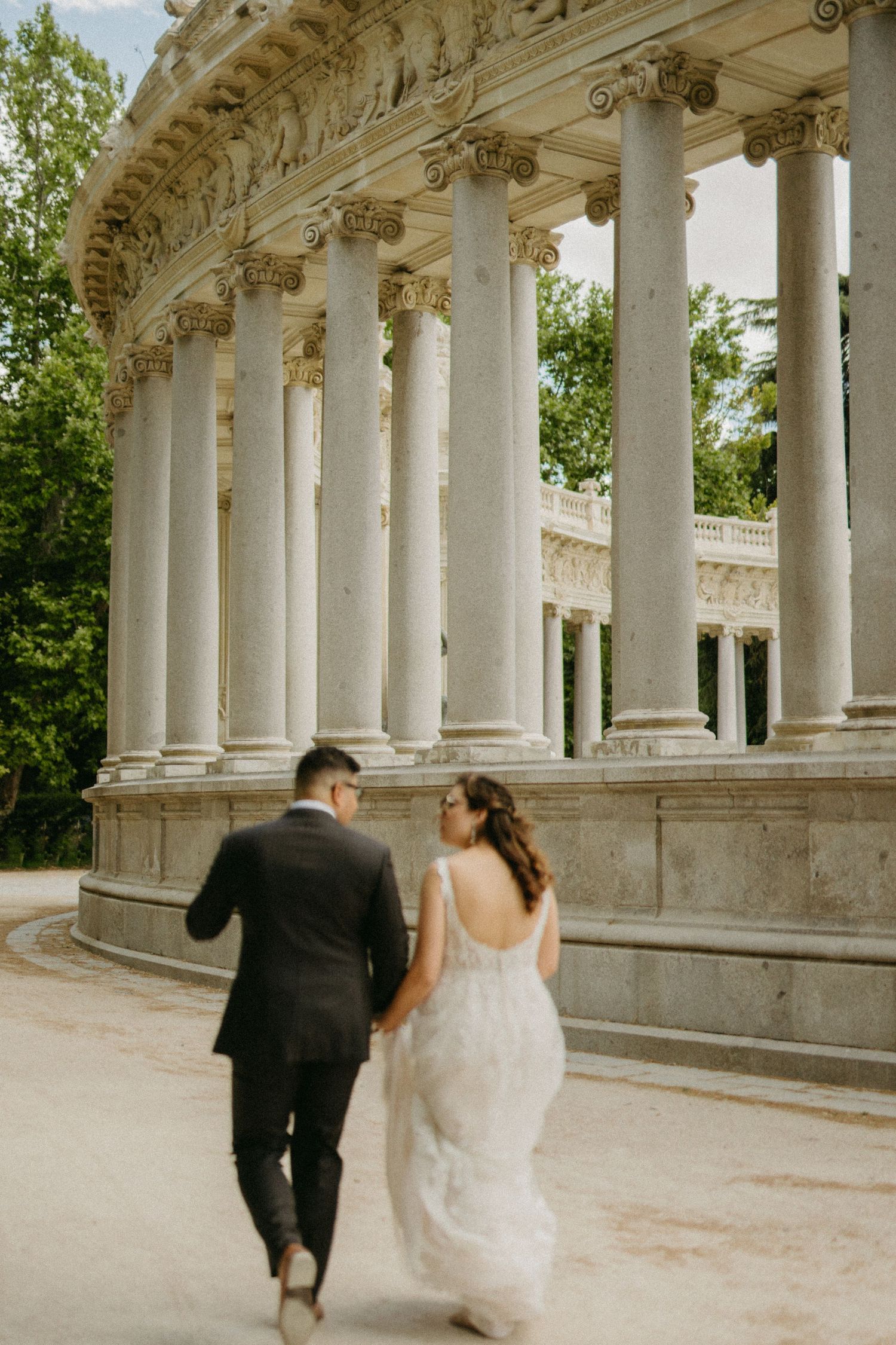 Bride and groom walk hand in hand past elegant white classical columns in a sunlit park setting.