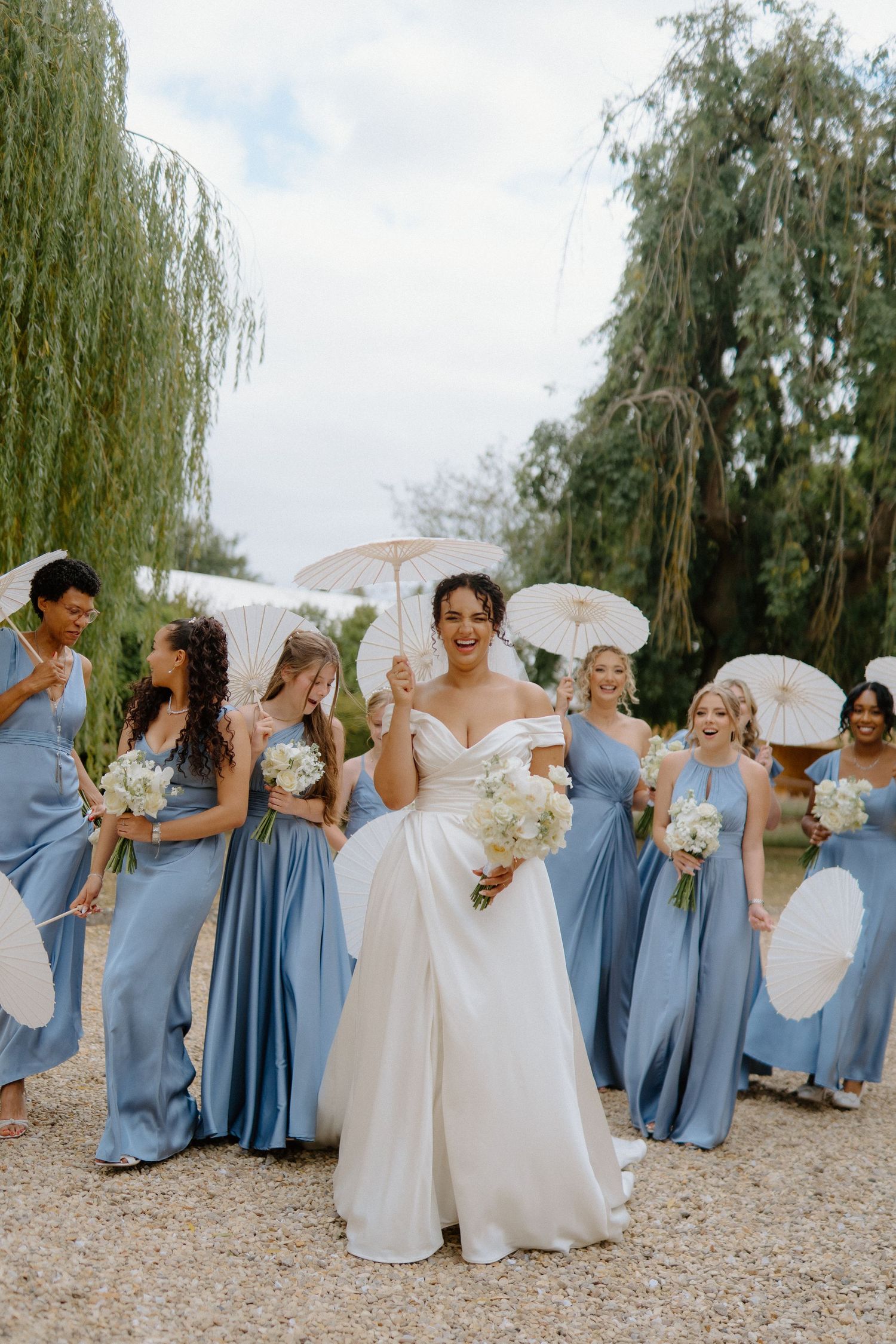Smiling bride in white gown leads bridesmaids in blue dresses holding parasols under willow trees.
